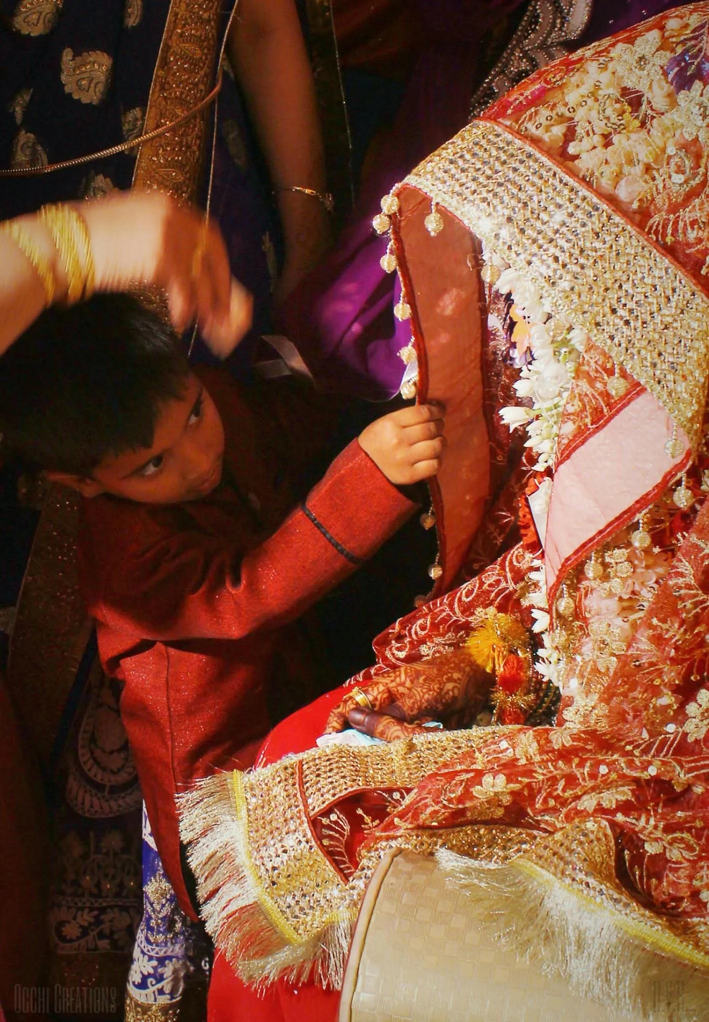 A young boy in traditional attire leaning on elaborately decorated wedding attire, with women assisting in the background.