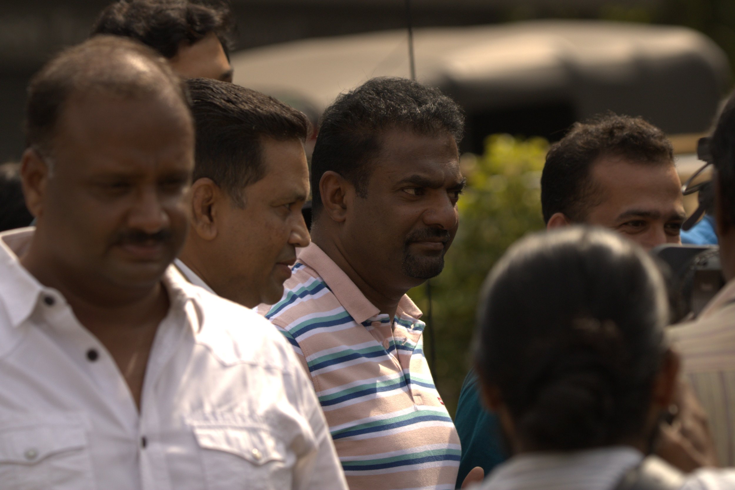A group of people standing close together outdoors, including a man in a striped shirt, a man in a white shirt, and several others, with a blurred vehicle and greenery in the background.