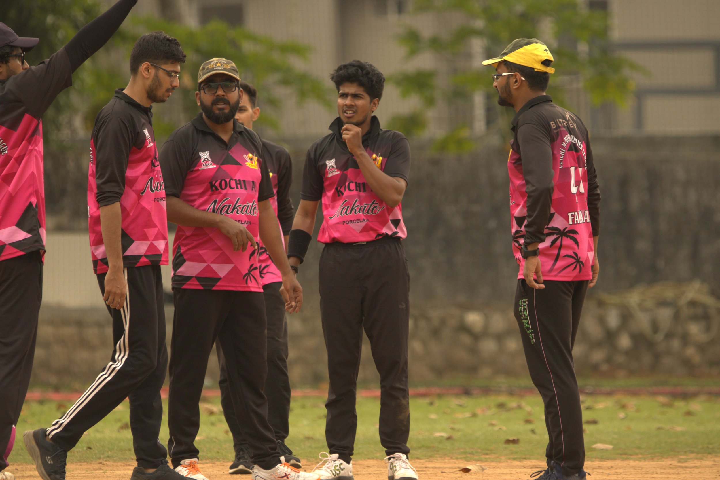 Group of cricket players in black and pink uniforms on a field, some wearing caps and sunglasses, engaged in a discussion during a game.