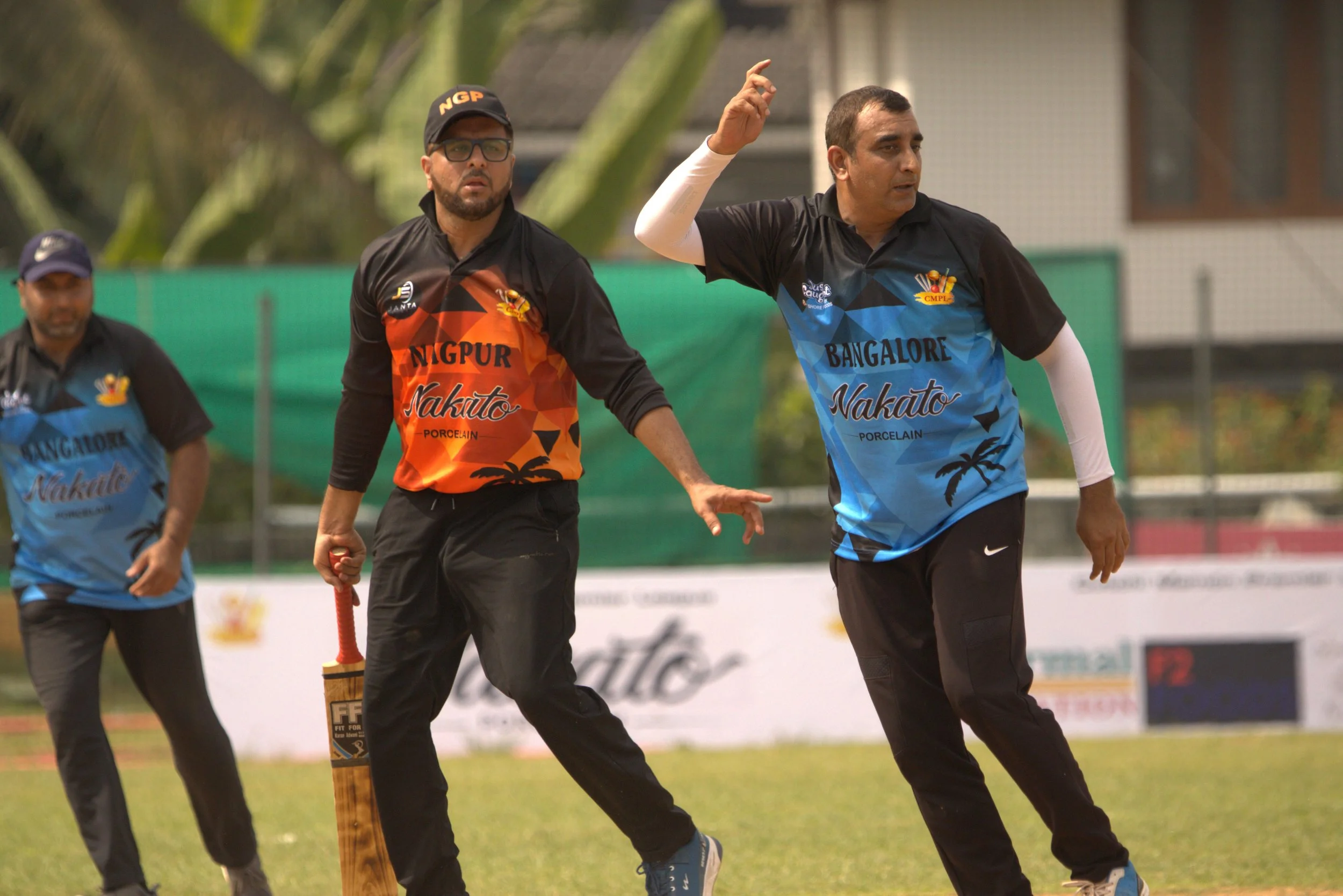 Three men playing cricket on a grassy field. Two are in focus, one in black and orange uniform, and the other in black and blue uniform, with one of them holding a cricket bat and the other gesturing with his hand.