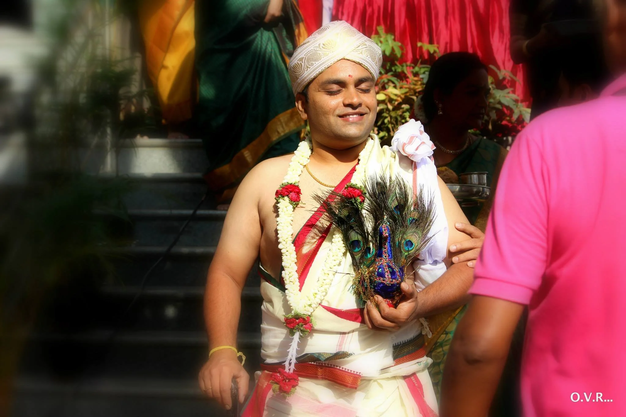 Man dressed in traditional Hindu ceremonial attire holding peacock feathers, wearing garlands, and standing in front of a group of women during a religious or cultural ceremony.
