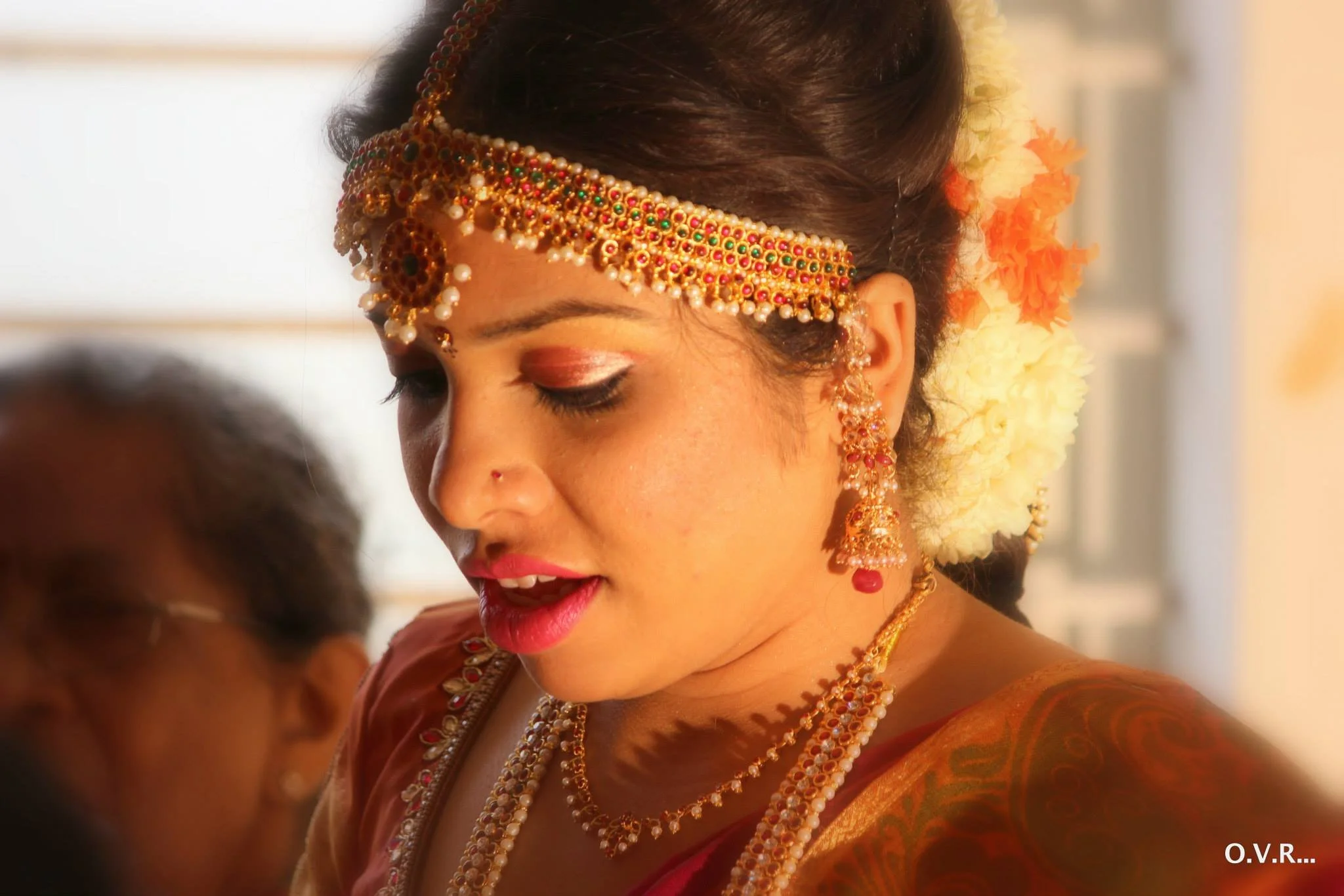 A woman dressed in traditional Indian wedding attire with intricate gold and red jewelry, floral hair accessories, and colorful saree, looking down.