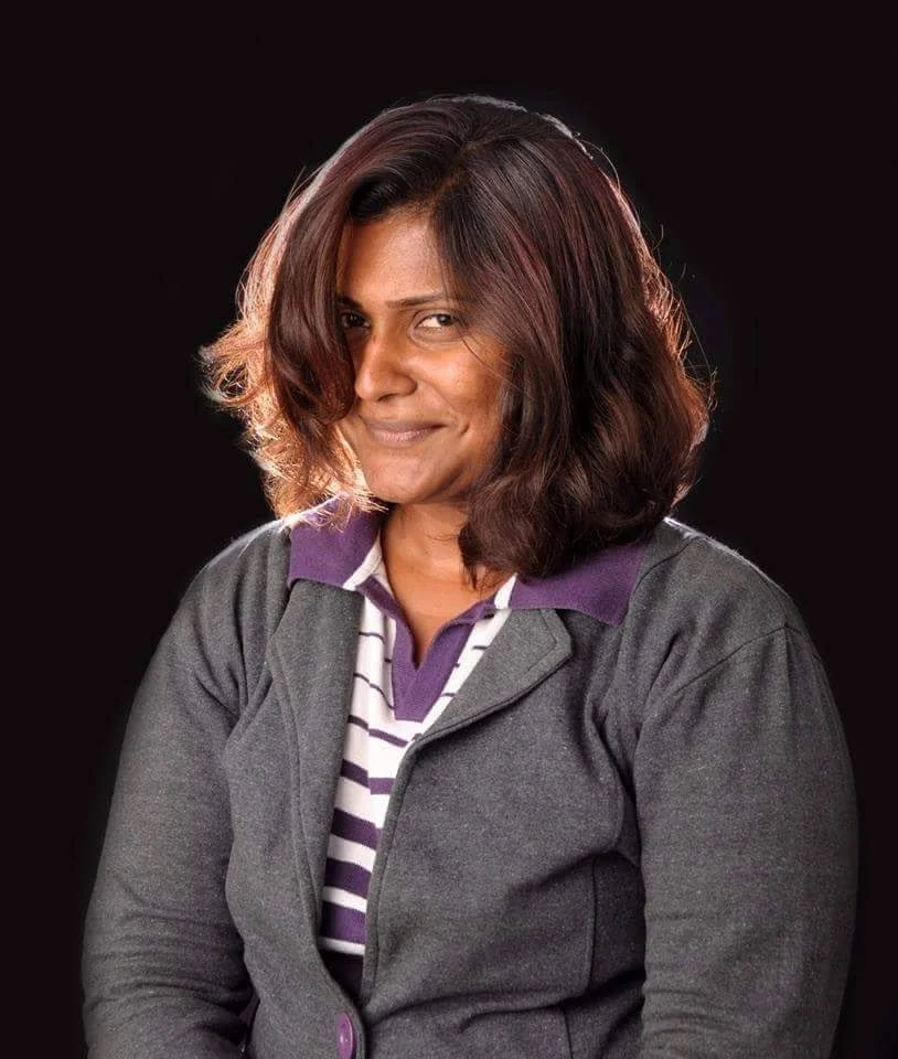 Portrait of a woman with shoulder-length wavy hair, wearing a gray jacket over a striped shirt, smiling slightly against a black background.