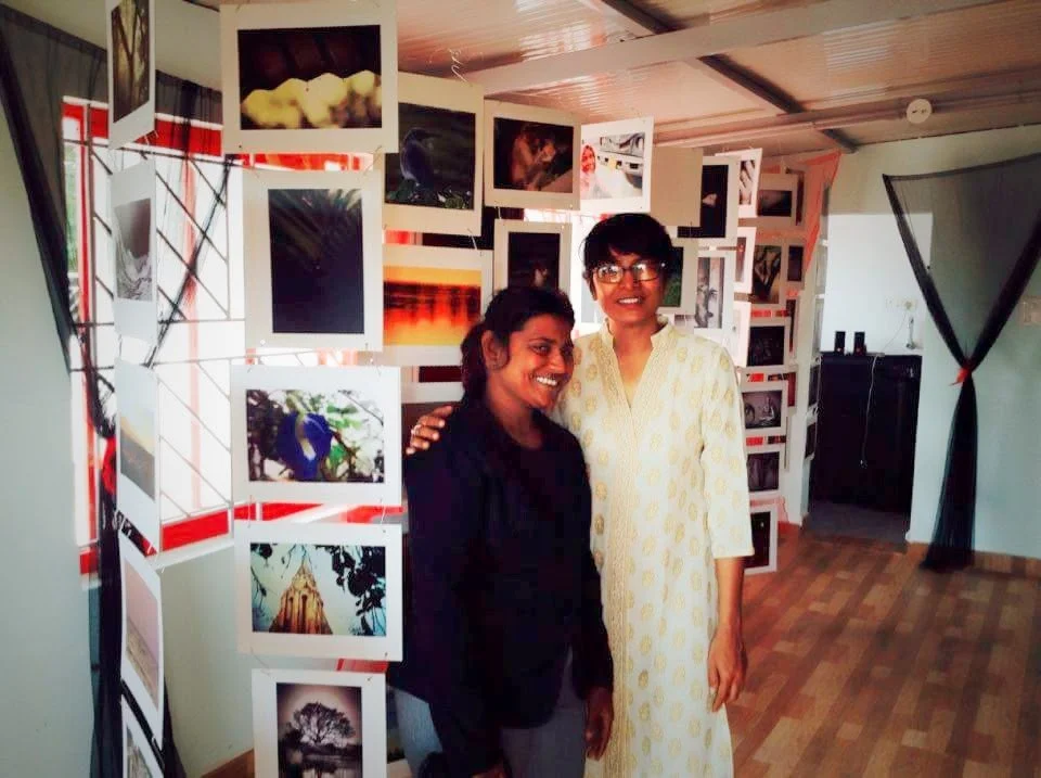 Two women smiling, standing together in front of a photo display in an indoor gallery setting.