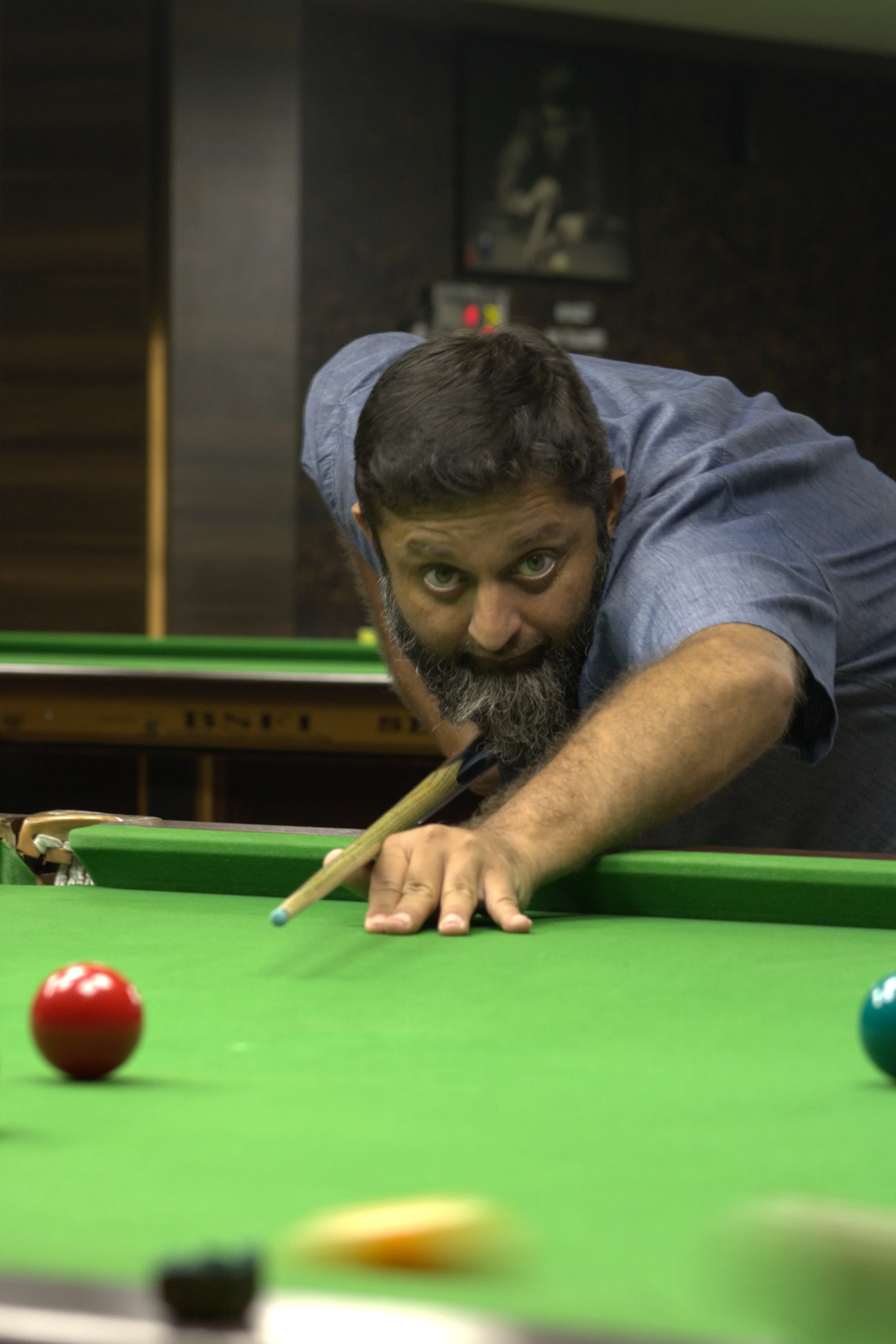 A man with a beard and blue shirt playing snooker, aiming to hit the cue ball on the green pool table with a cue stick.