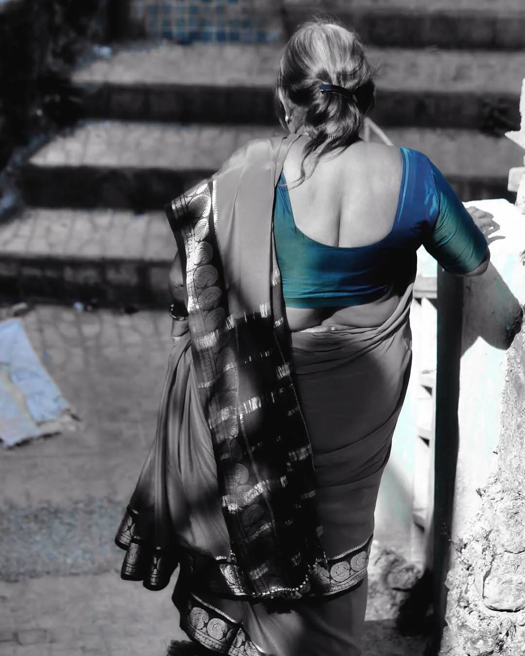 A woman with long hair tied back, wearing a blue and green blouse and a patterned sari, leaning against a stone railing with her back to the camera.
