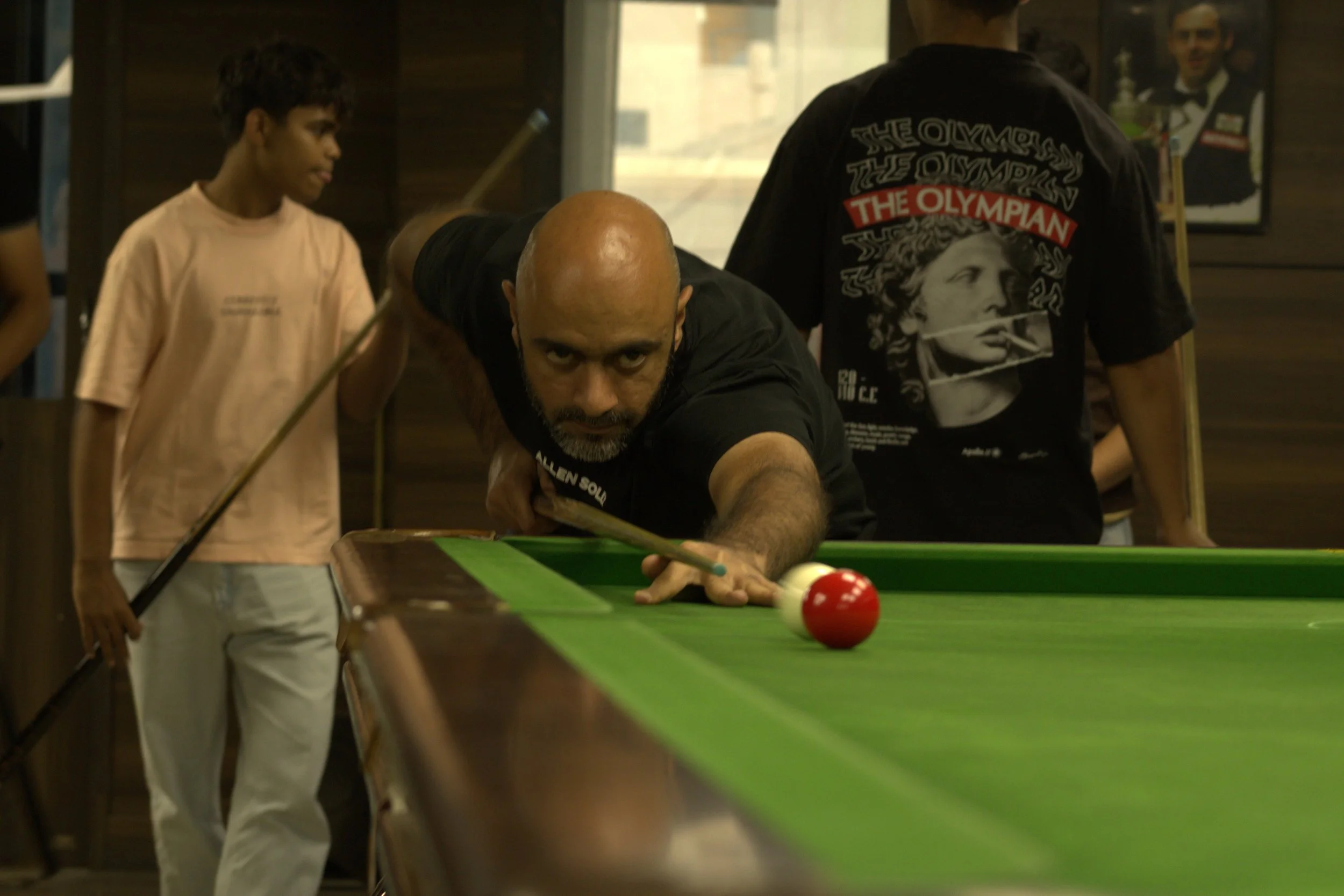 A man with a shaved head and beard is taking a shot in a pool game, leaning over the green table and aiming at the red and white cue ball. A boy in a light-colored shirt holds a pool cue in the background, observing. Other people are visible in the b