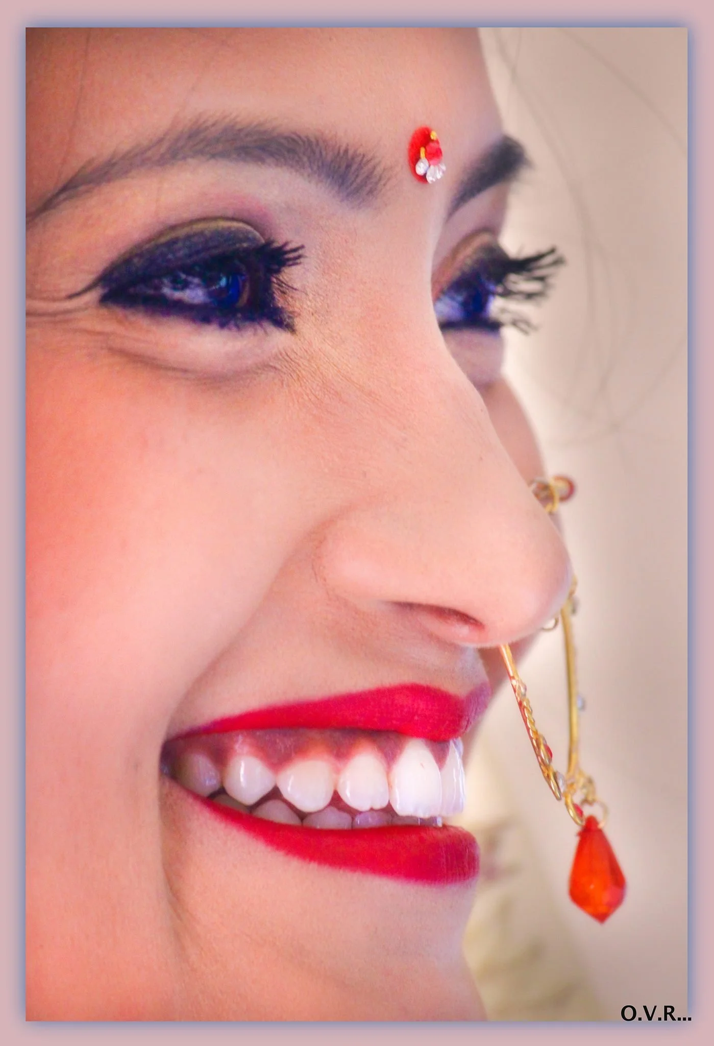 Close-up of a woman's face showing her eyes, nose, and mouth, with decorative jewelry, bright red lipstick, and makeup. She has a small red bindi on her forehead with embellishments, and a nose chain with a red gemstone hanging from it.