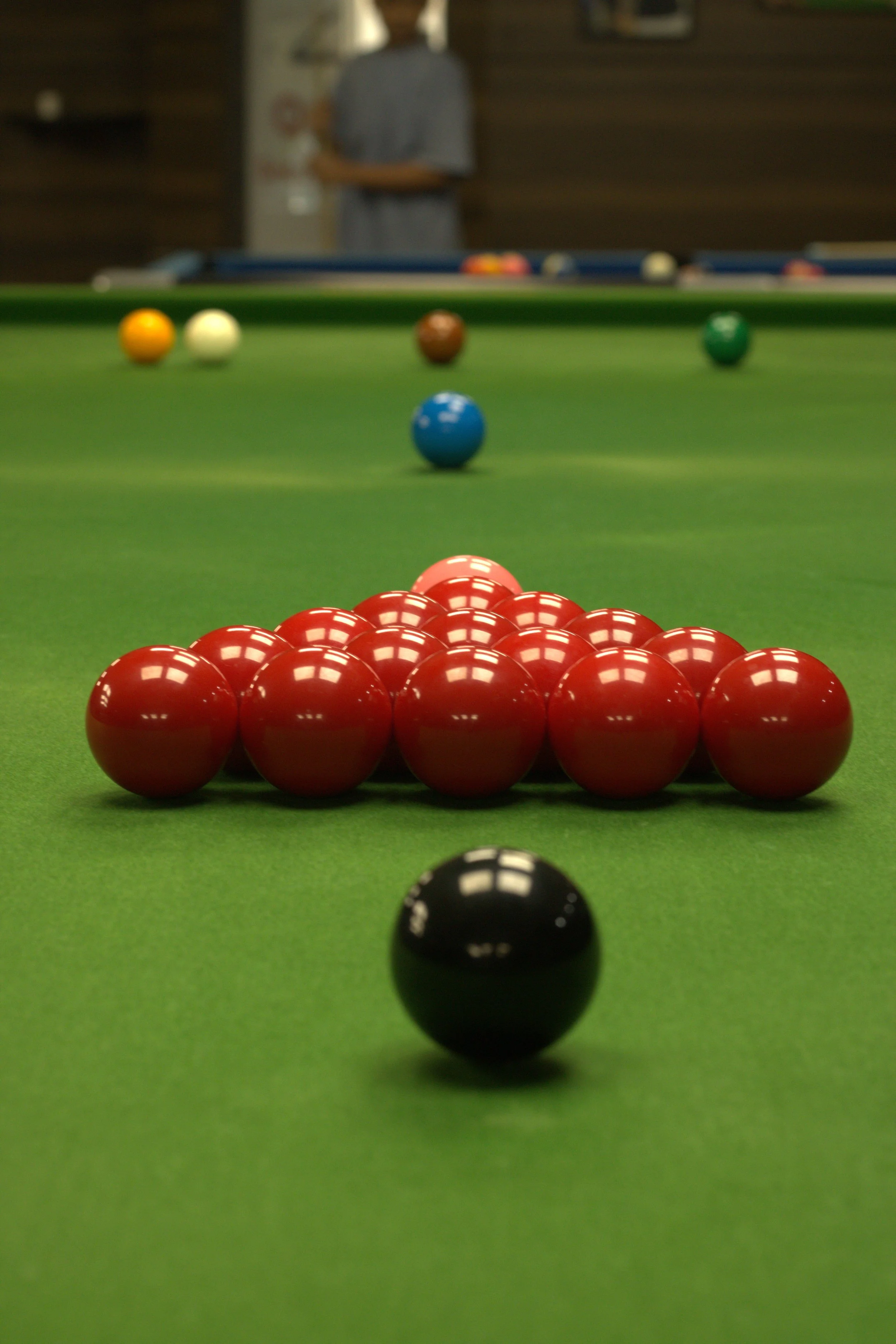 A game of billiards with red balls arranged in a triangle, a black cue ball in the foreground, and other colored balls scattered on the green felt table in the background, with a person standing near the table.