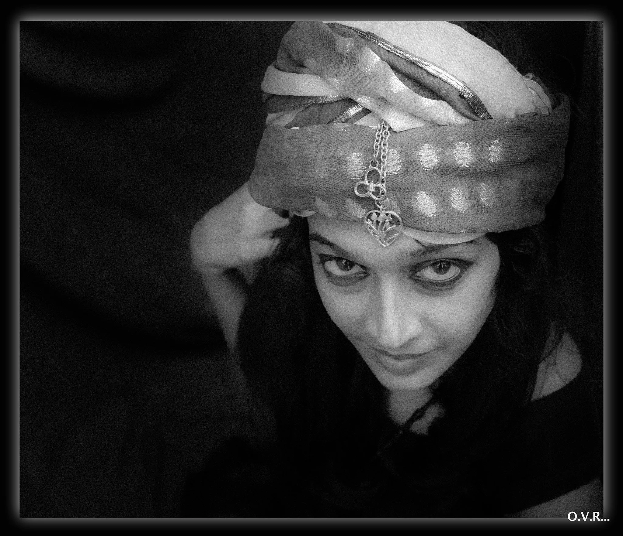Woman with dark hair wearing a headwrap with a decorative chain and pendant, looking up at the camera, photographed in black and white.