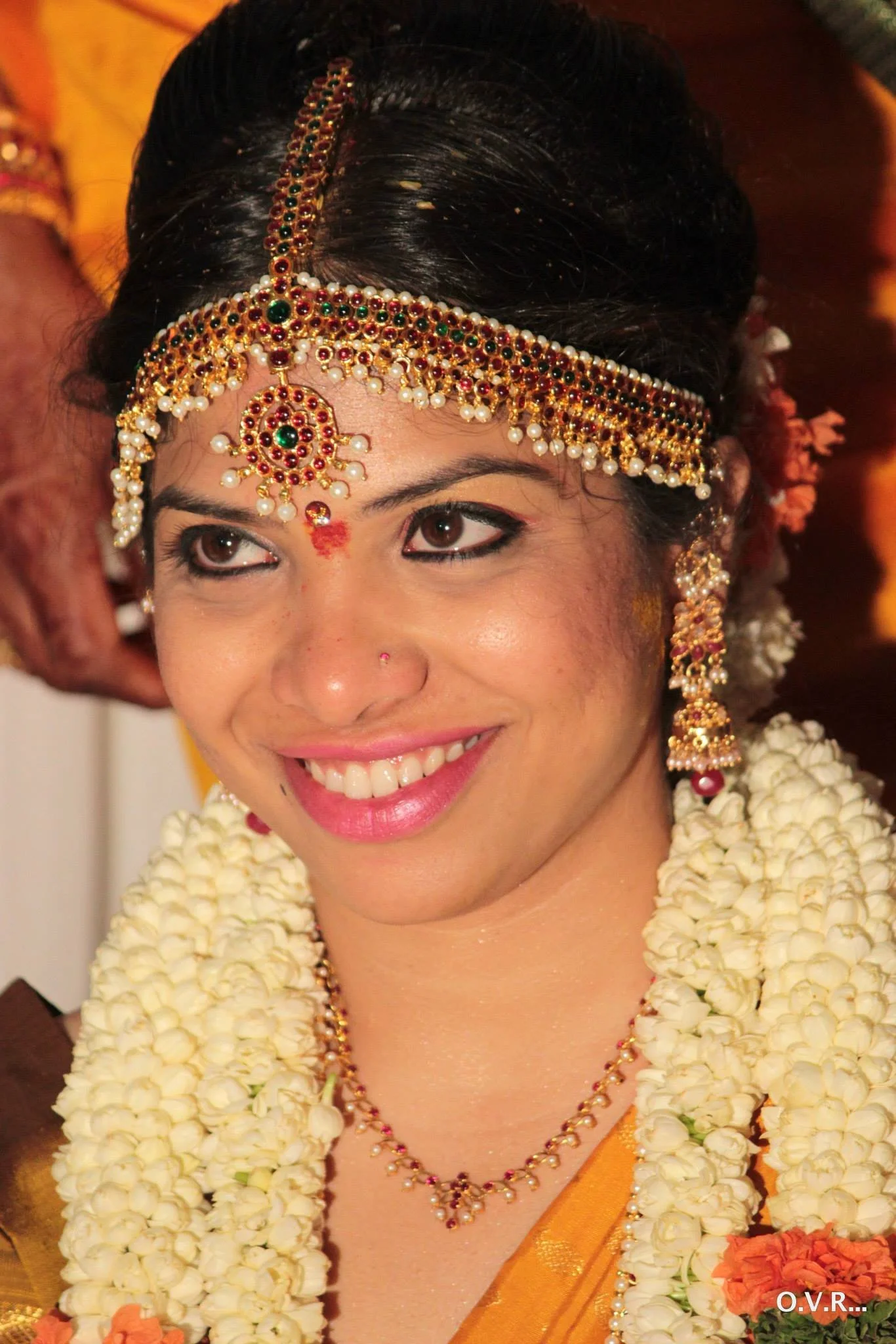 A woman dressed in traditional Indian wedding attire with jewelry, makeup, and flowers, smiling during a wedding ceremony.