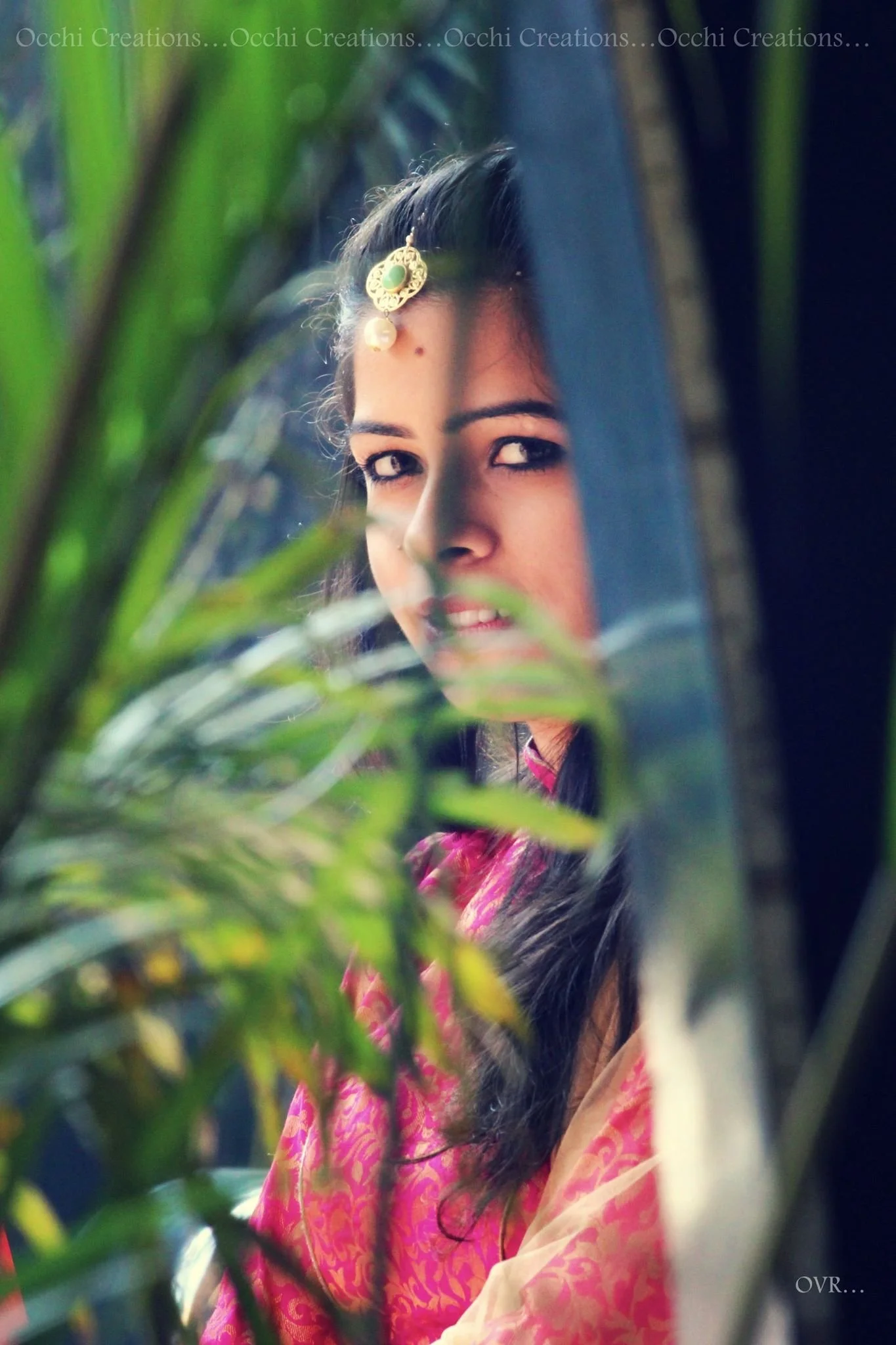 A young woman in traditional Indian attire peeking through green foliage, with her face partially obscured by leaves.