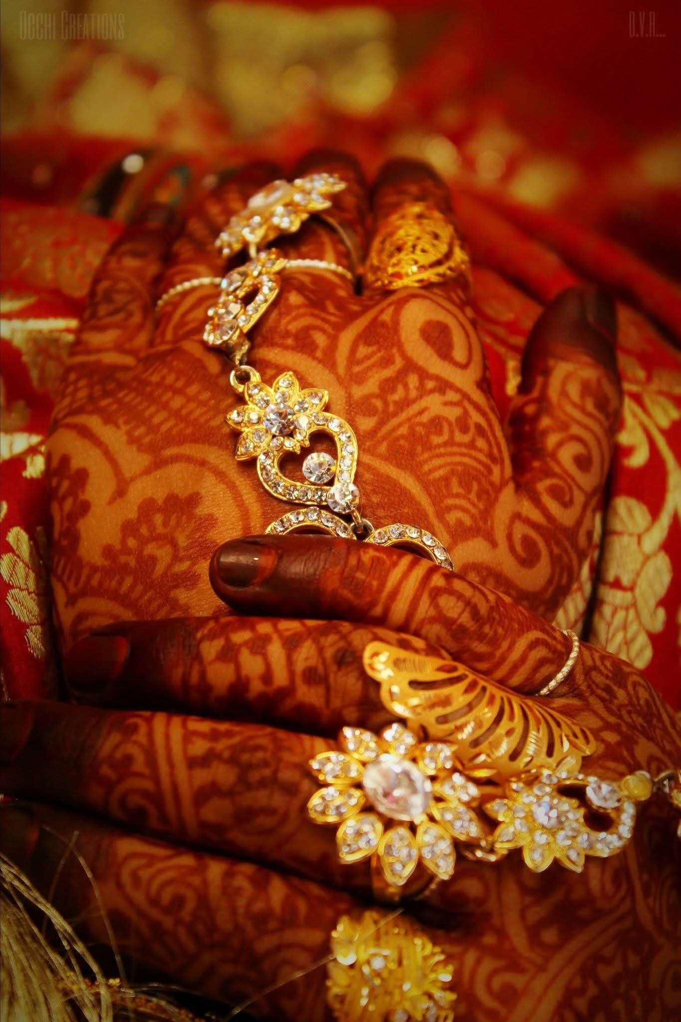 Close-up of a hand decorated with henna, adorned with gold jewelry including rings, a bracelet, and a necklace with floral and heart-shaped designs, resting on richly patterned red and gold fabric.