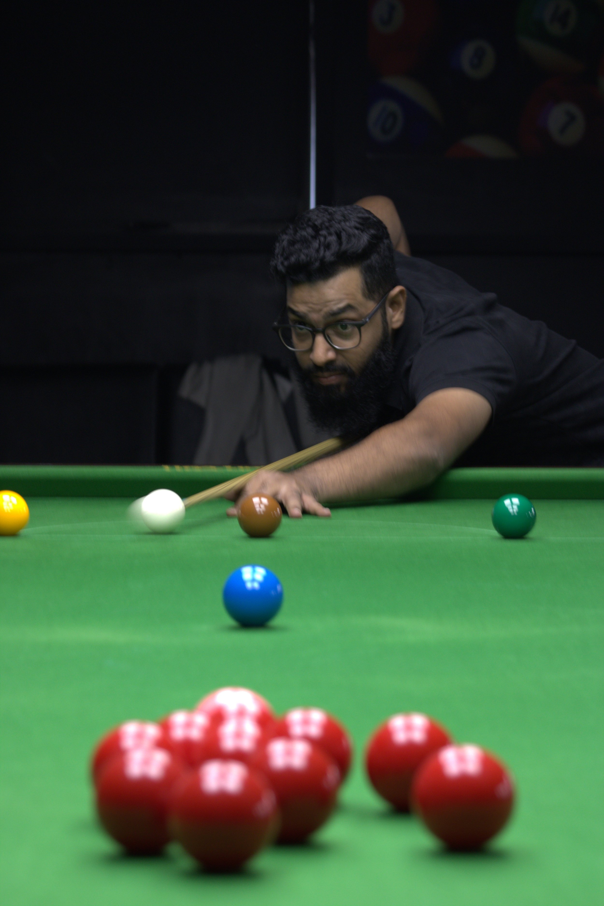 A man with glasses and a beard playing pool, aiming to hit a white cue ball near the brown ball, with multicolored balls in the background and red balls in the foreground.