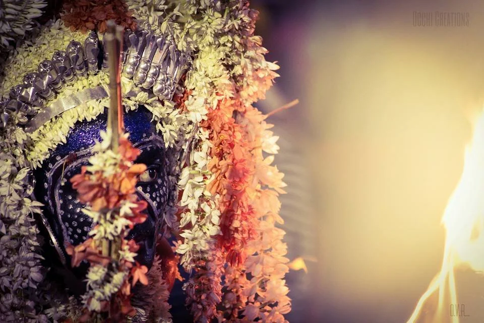 A person dressed as a traditional Indian deity or goddess, adorned with an elaborate floral headdress and jewelry, holding a lit torch, with a fire on the right side.