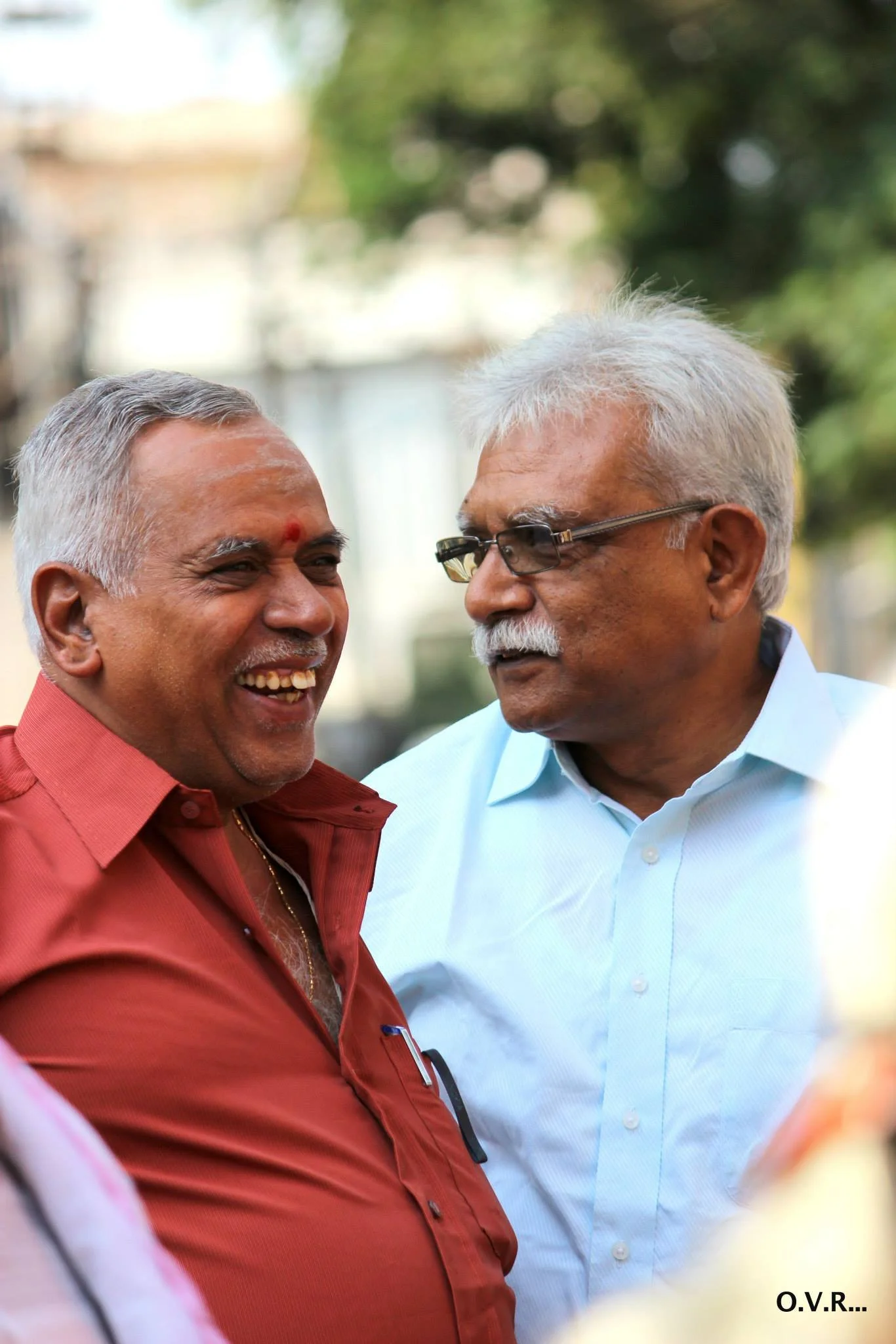 Two smiling older men having a conversation outdoors, one wearing a red shirt with a gold chain and the other wearing glasses and a light blue shirt, with blurred trees in the background.