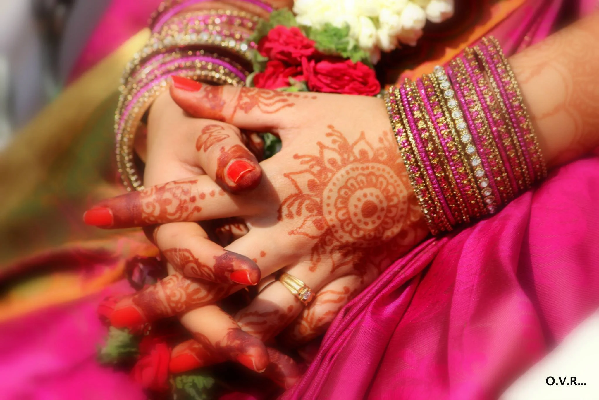 Close-up of a bride's hands decorated with intricate henna designs, wearing traditional pink and gold jewelry and a pink sari, holding a bouquet of red and white flowers.
