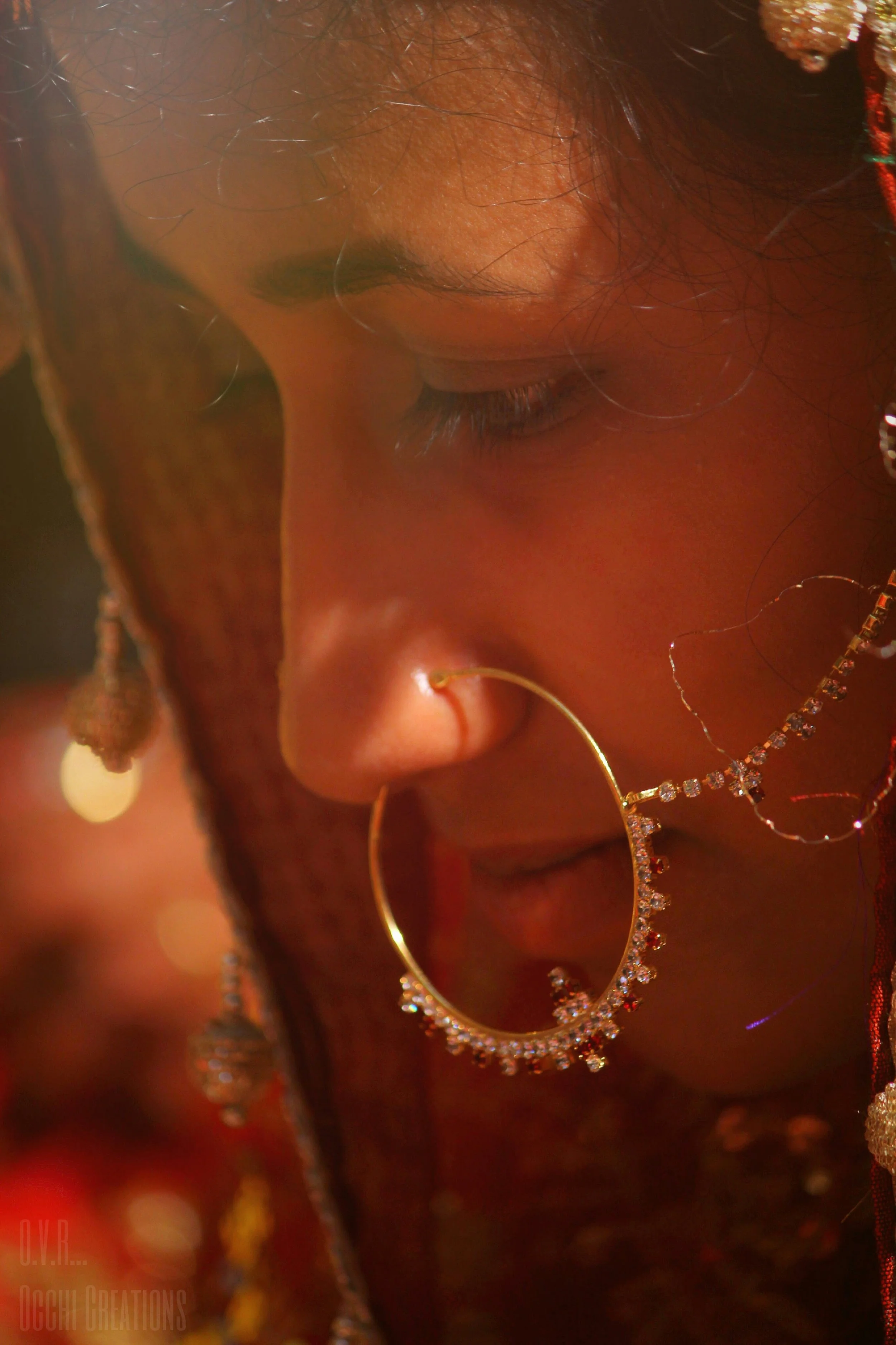 A woman with traditional hoop earrings and jewelry, posed with her head tilted downward, partially obscuring her face, which is close-up and detailed.