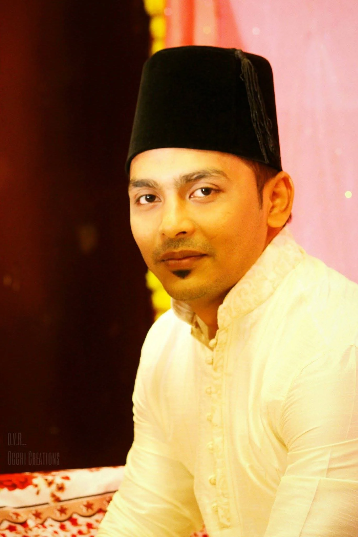 A man wearing traditional Indian attire and a black top hat, sitting indoors with a pink and yellow backdrop.