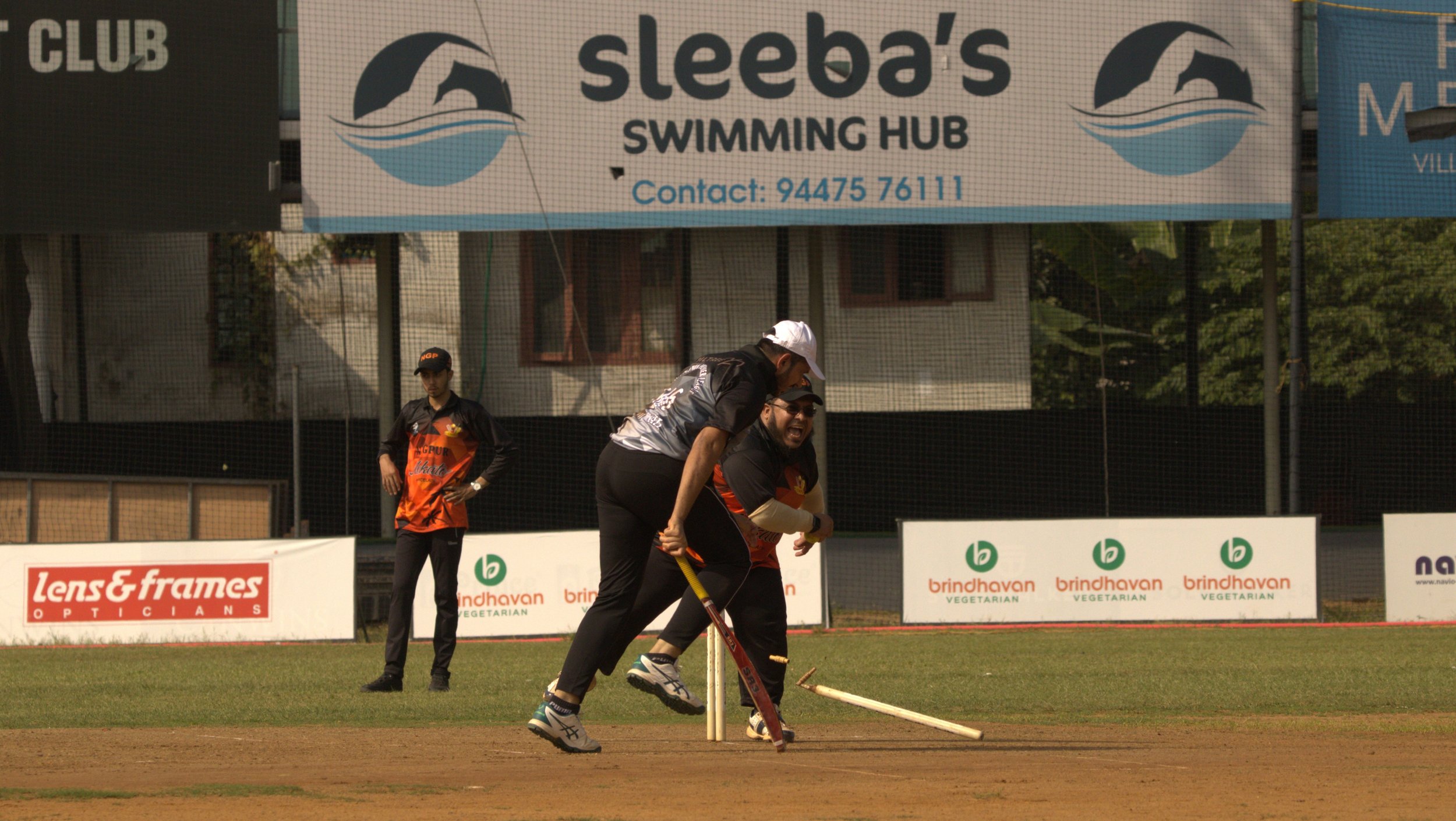 Two cricket players celebrate on the field near the stumps with a third person watching nearby. Advertisement banners are visible in the background.