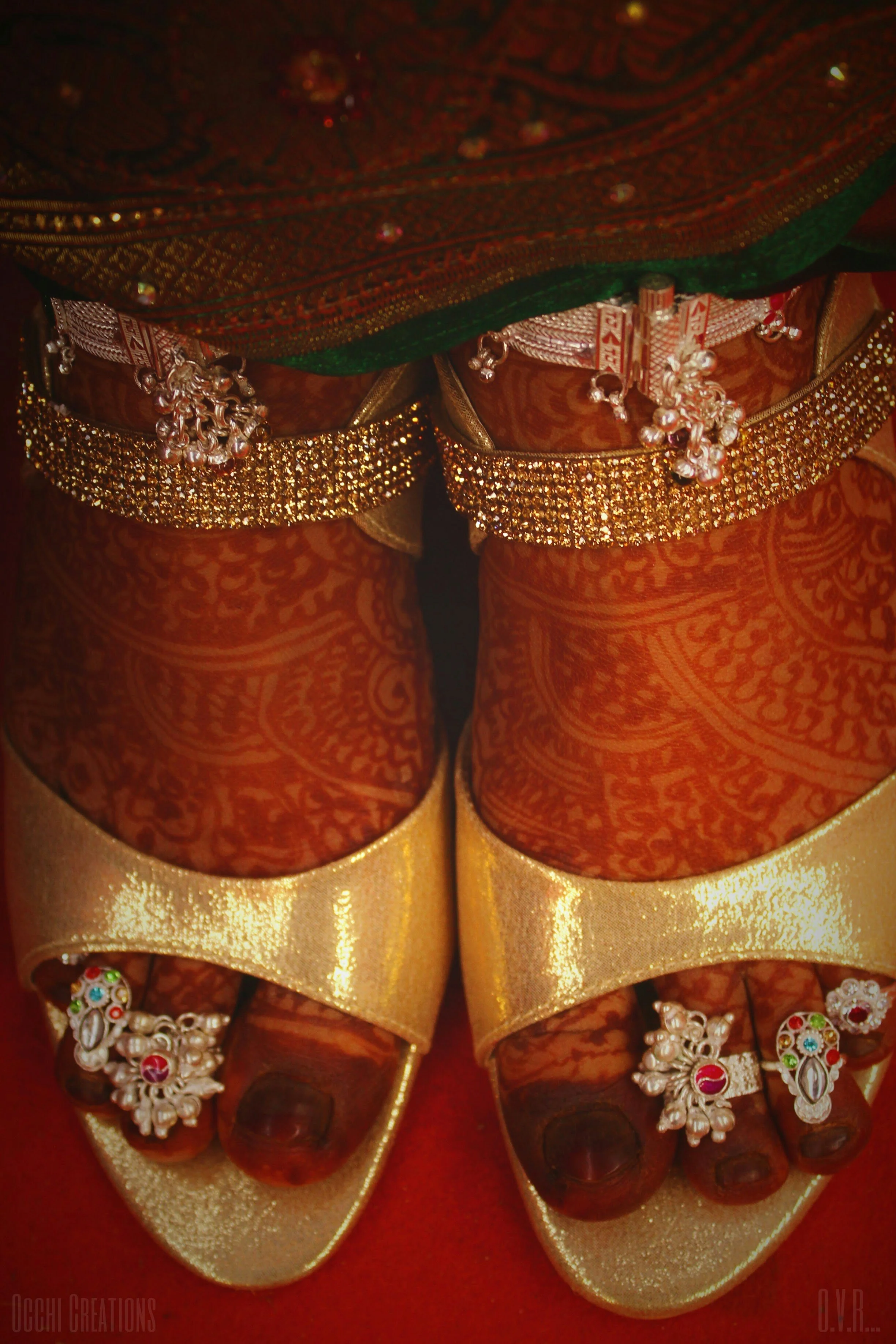 Close-up of feet adorned with traditional Indian jewelry, henna designs, gold sandals with charms, and ankle bracelets, indicating a festive or ceremonial occasion.