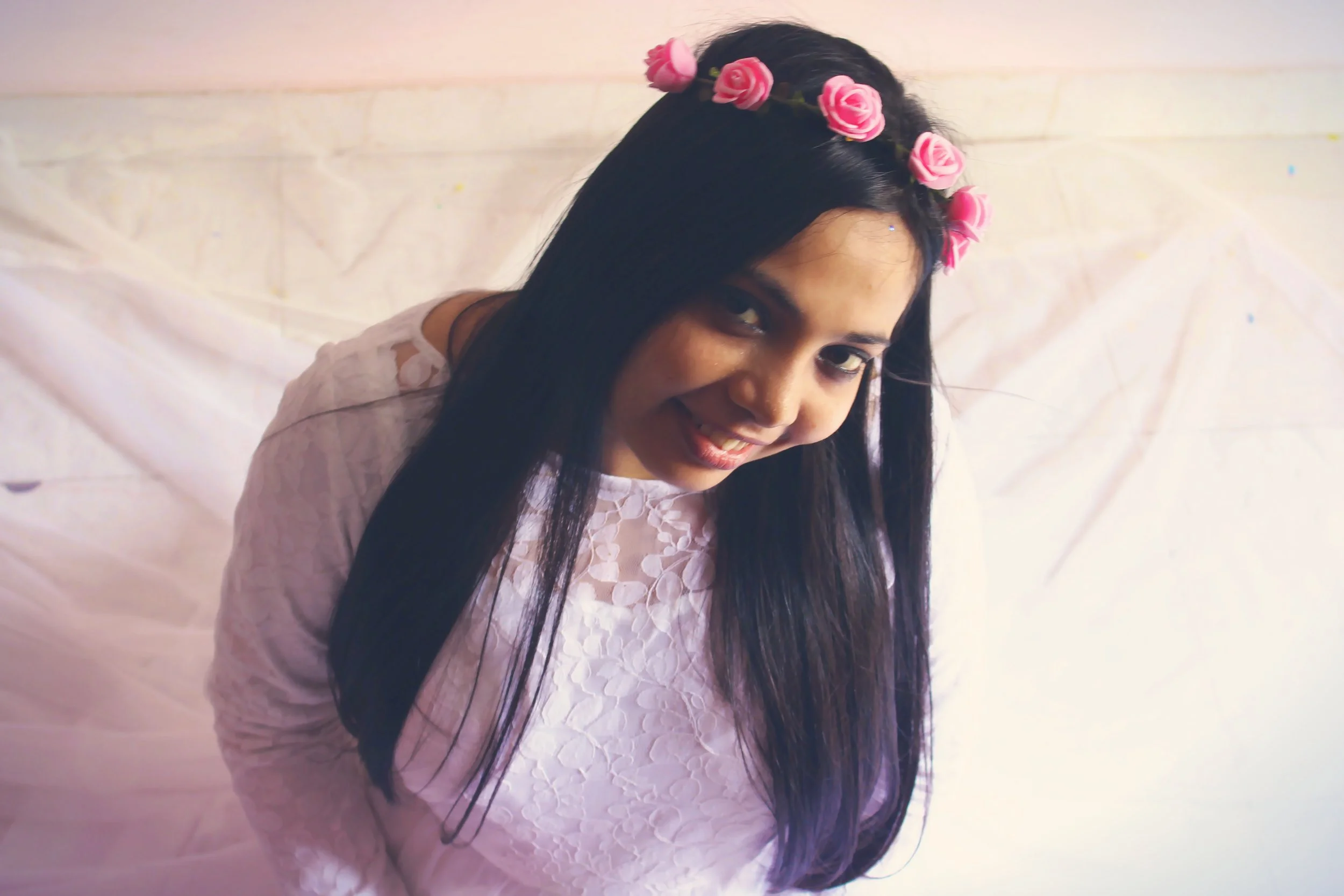 A young woman with long dark hair wearing a white lace dress and a pink rose flower crown, smiling and leaning forward.