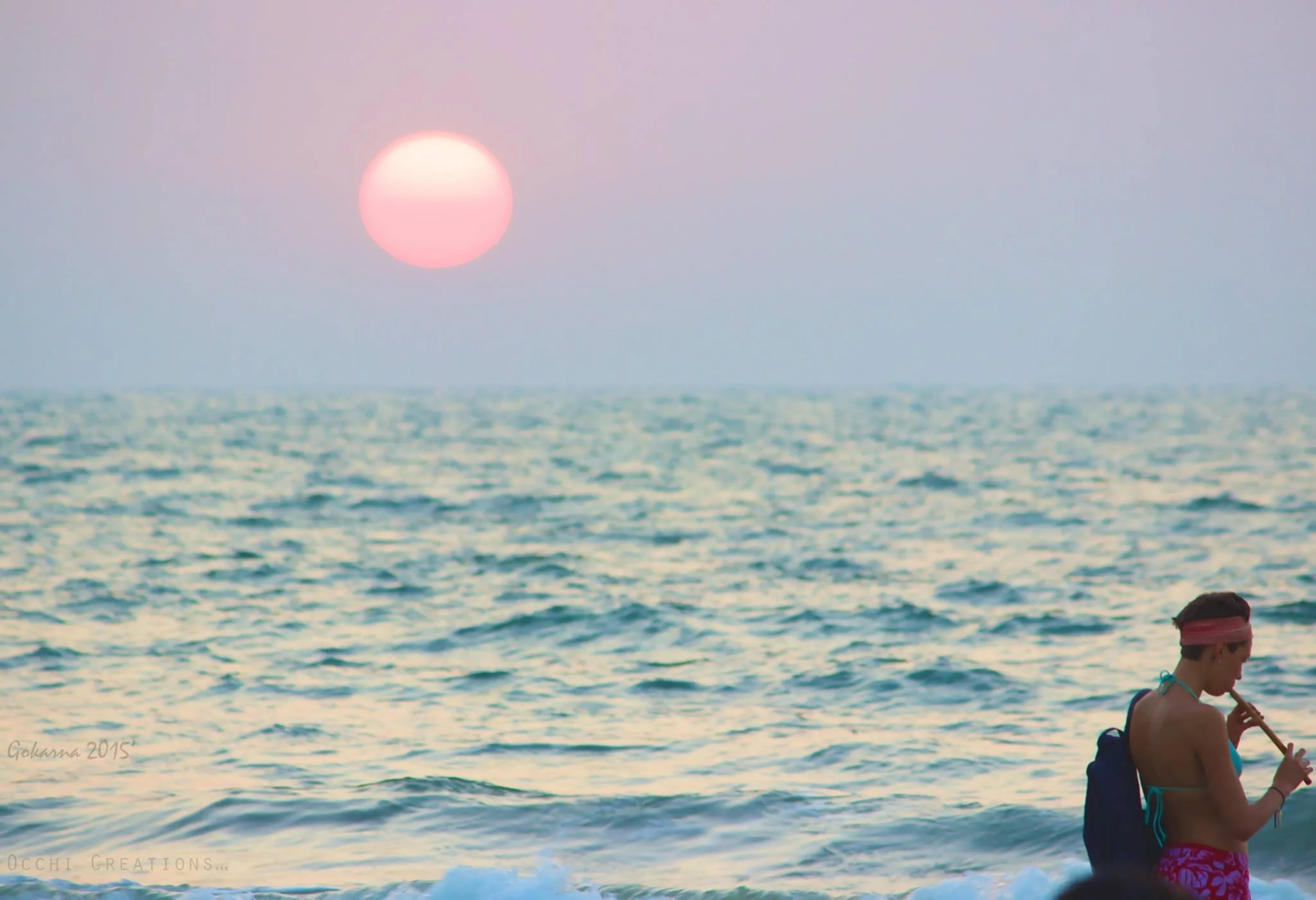 Person with a backpack playing a flute on the beach at sunset with pink and purple sky, large sun, and ocean waves.