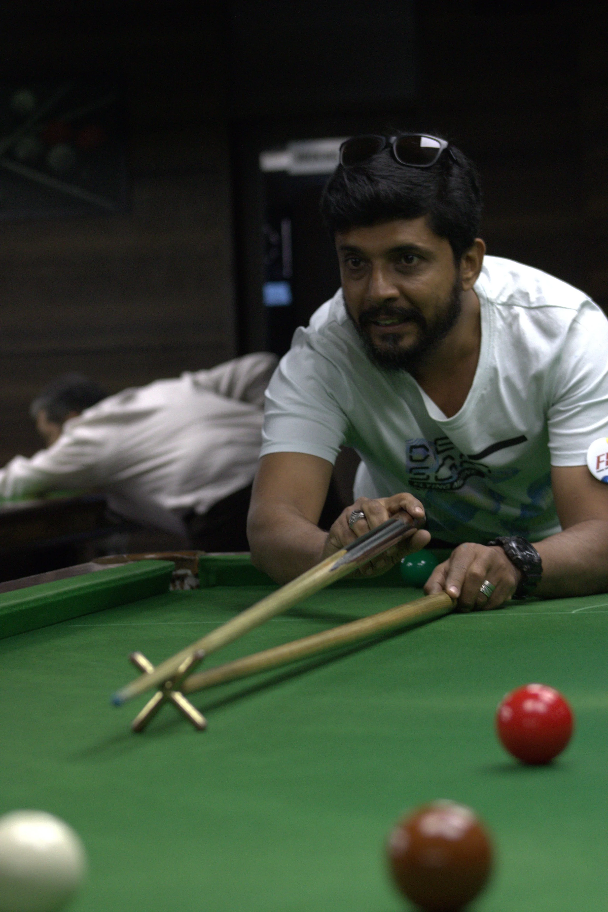 Man playing billiards, leaning over the pool table with a cue stick, focused on the game, wearing a white T-shirt, sunglasses on head, dark hair and beard, with other players in the background.