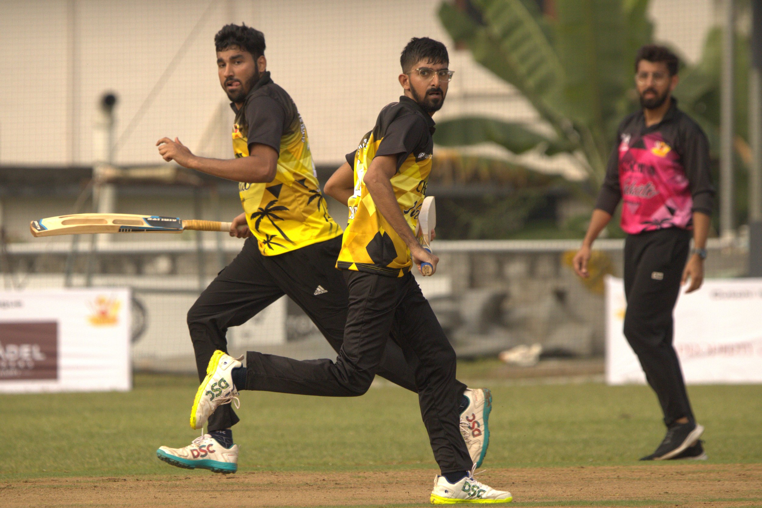 Two cricket players in yellow and black uniforms running on the field with a cricket bat, while a third player in a pink and black uniform stands in the background.