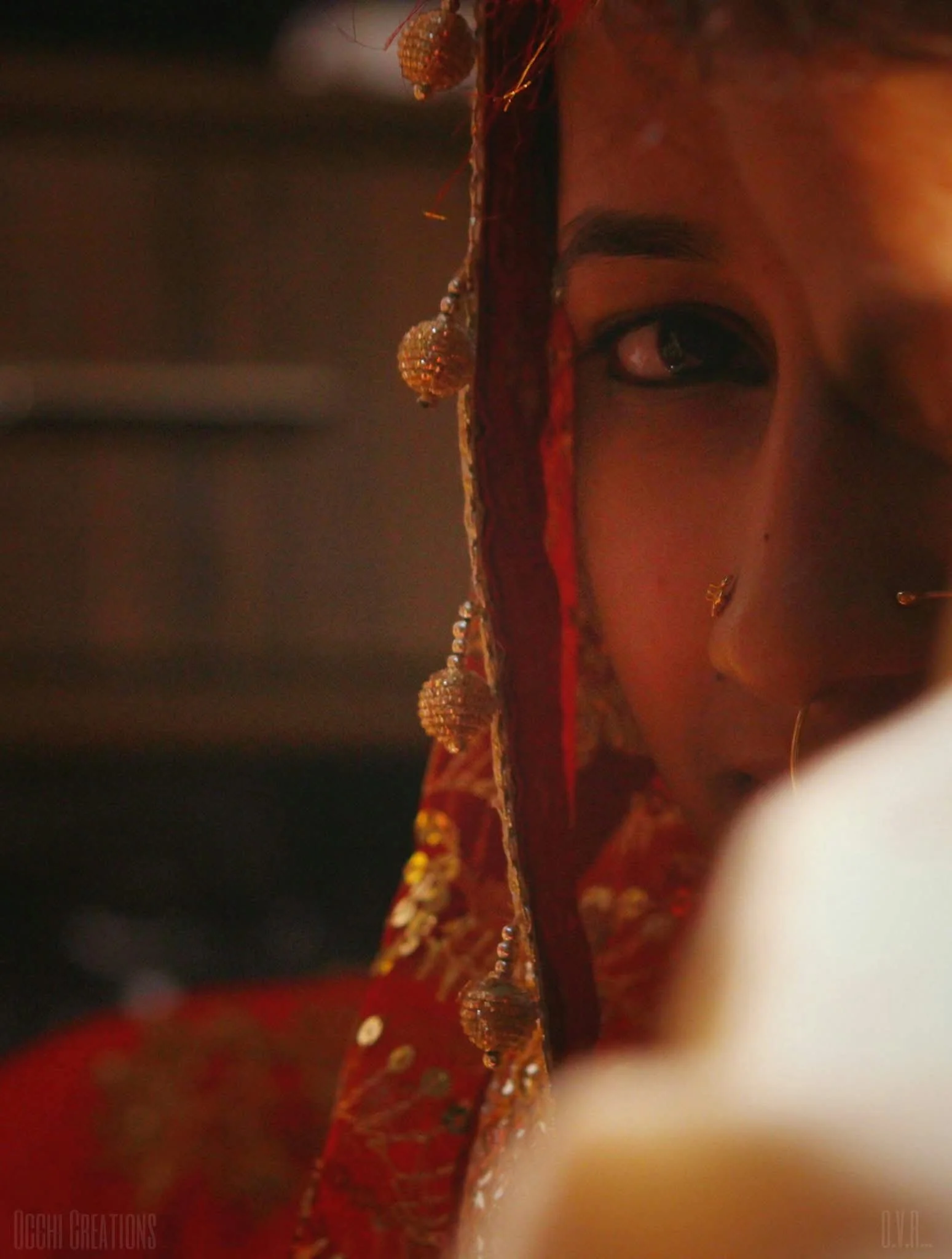 A woman dressed in traditional Indian attire with jewelry, partially covered by a red veil, looking over her shoulder.