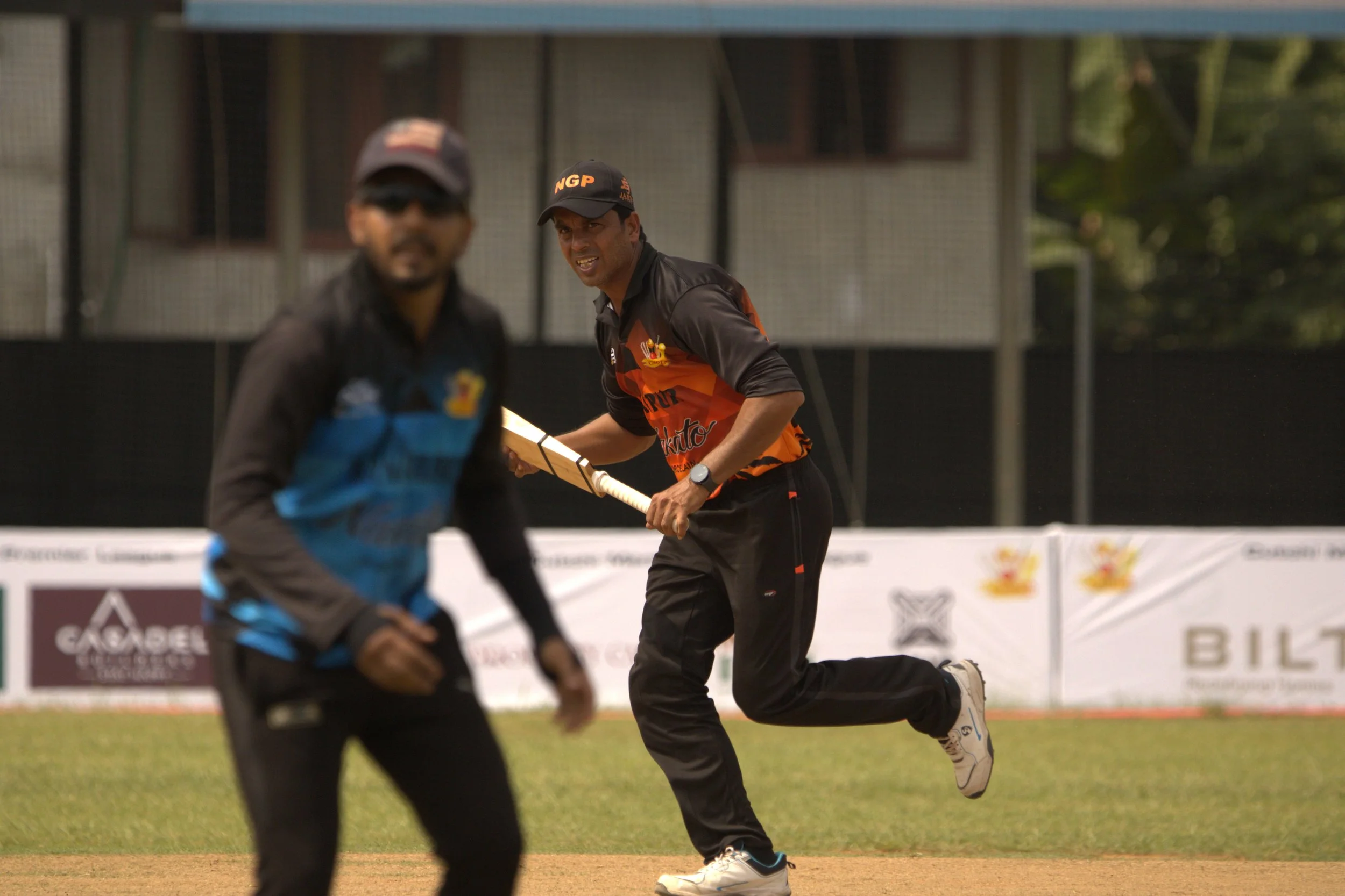 Two men playing cricket on a field, with one man in focus behind the other, holding a cricket bat and running, while the other man is slightly blurred in the foreground.