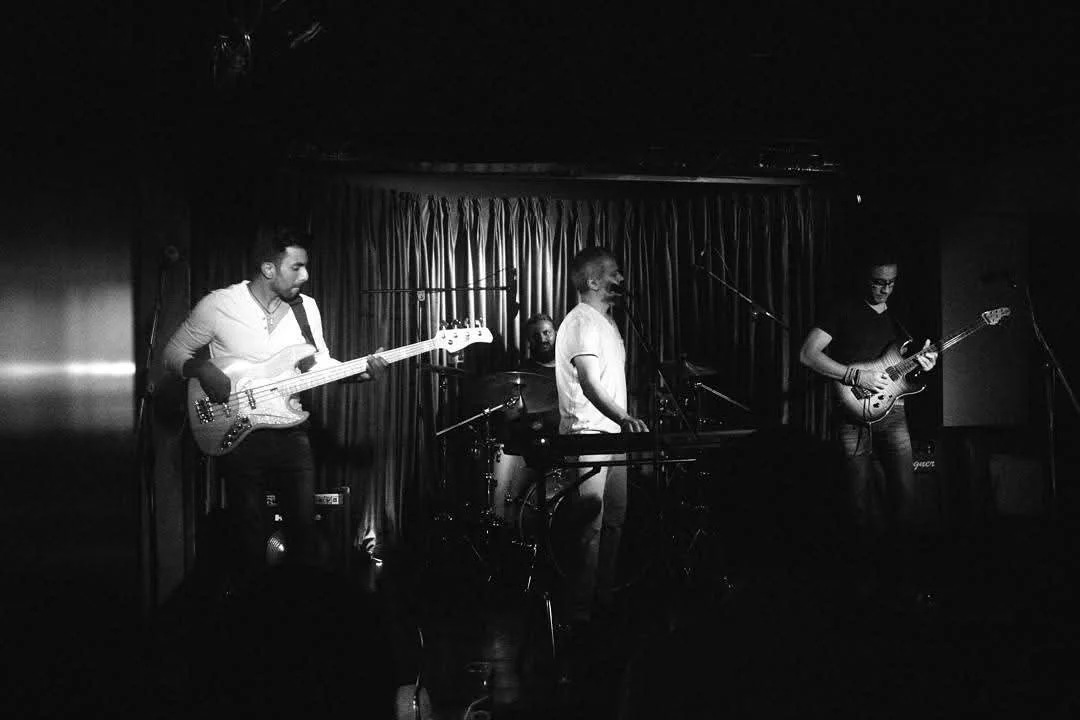 A black-and-white photo of a four-member band performing on stage with a curtain backdrop. The band includes a bassist, a drummer, a keyboardist, and a guitarist.