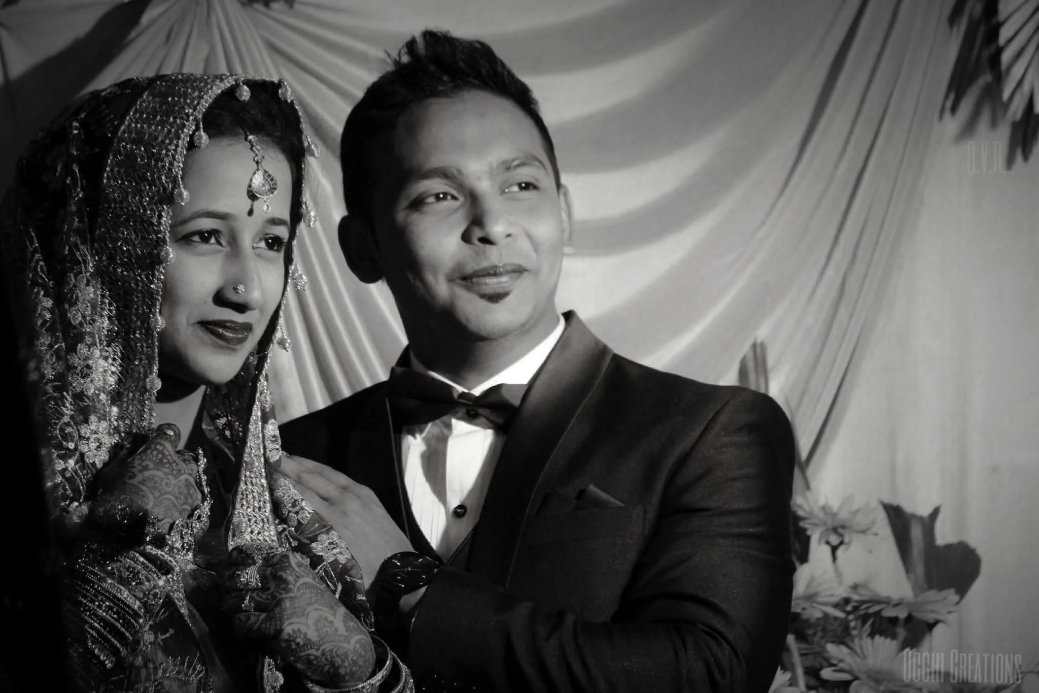 A black and white photo of a man in a tuxedo and a woman in traditional attire, standing close together at a wedding or special celebration event, with a decorative curtain and flowers in the background.