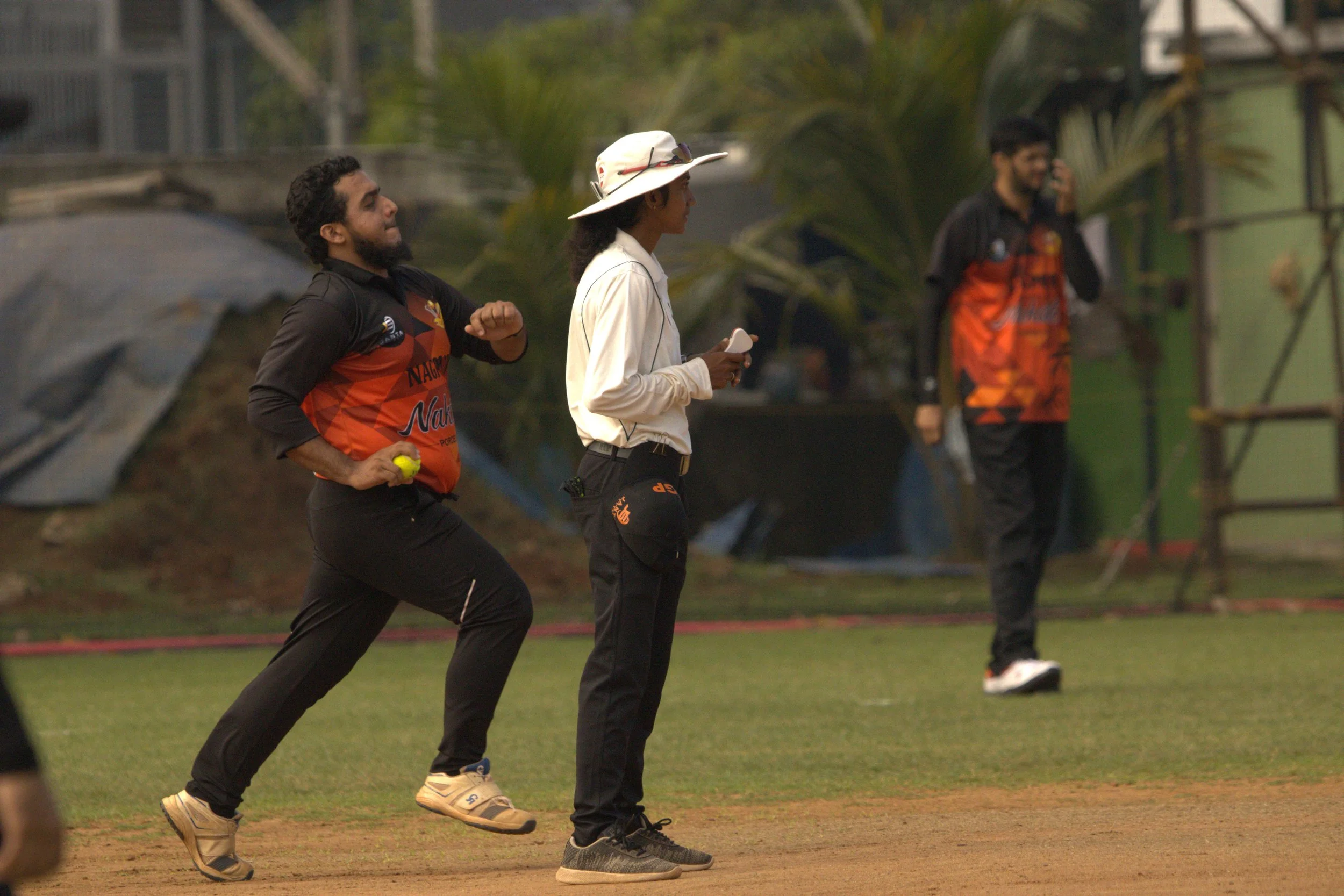 Two cricket players and a woman standing on the field, with one player holding a yellow cricket ball and in motion, and the woman wearing a hat and holding a clipboard, during a cricket match.