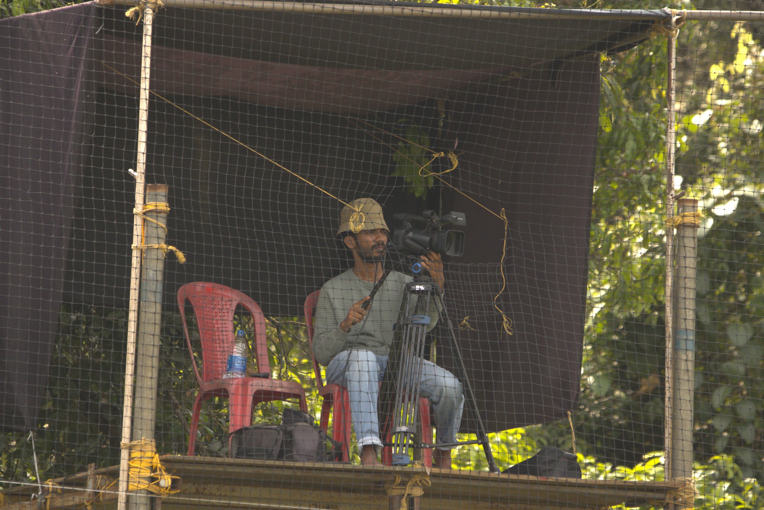 A man sitting on a red plastic chair on a wooden platform, operating a video camera. He is wearing a beige hat and gray clothing, with a bottle of water and a black bag nearby. The platform is enclosed with black netting, and there are trees in the b
