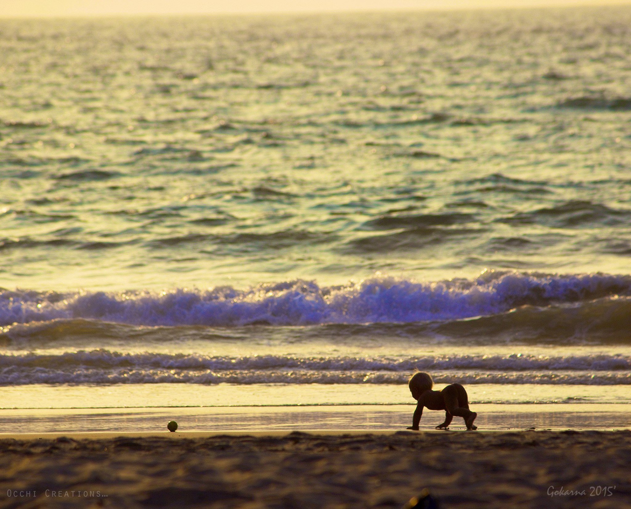 A young child playing on the beach during sunset, crawling near the shoreline with ocean waves in the background.