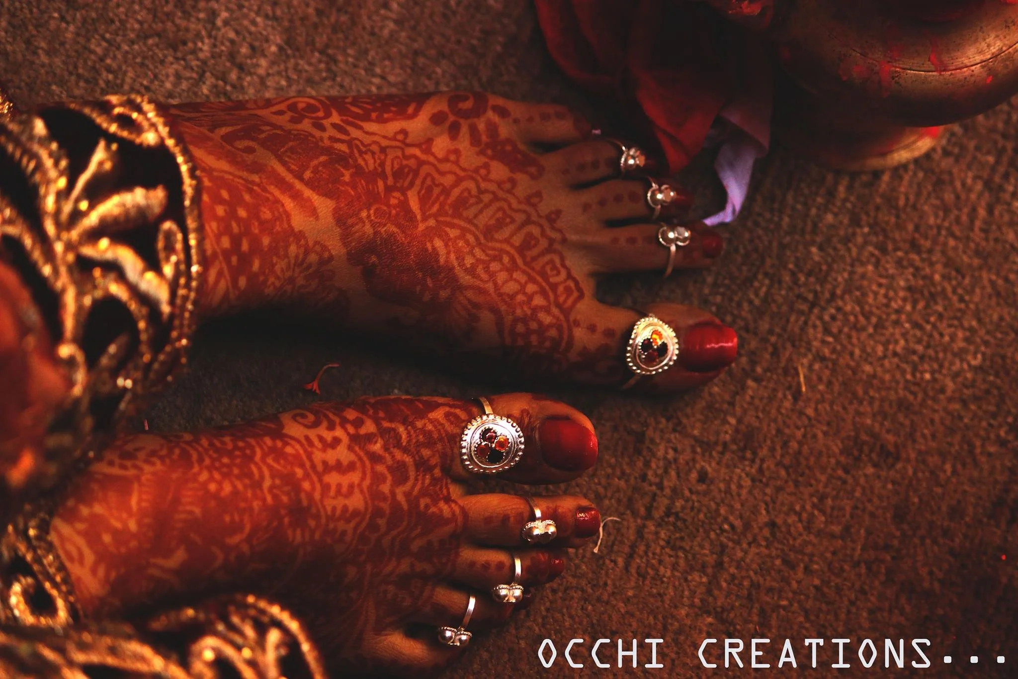Close-up of intricately decorated henna-covered hands adorned with rings and bangles, resting on a textured surface, with a copper vessel nearby.