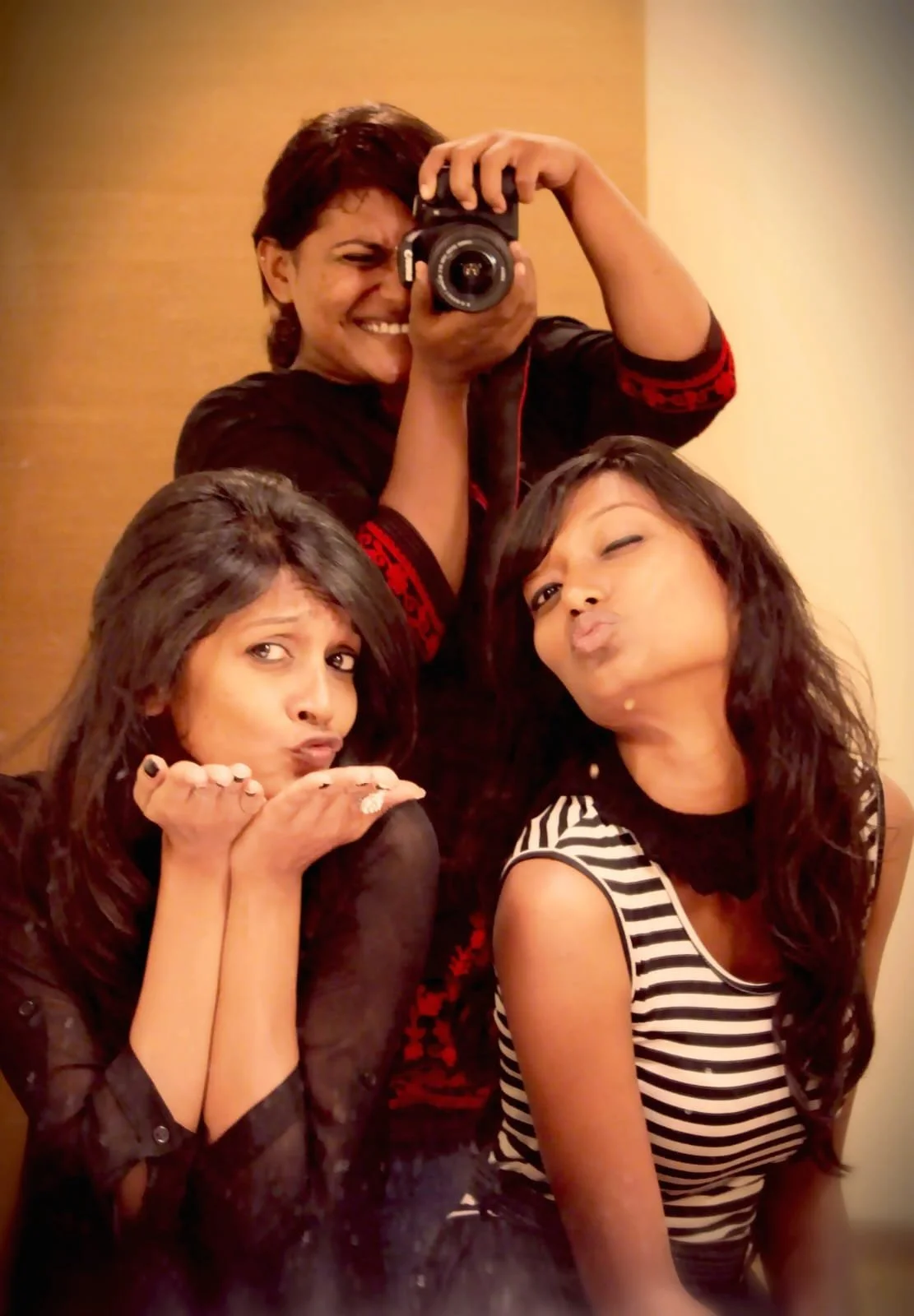 A woman with short dark hair takes a selfie with two women in front pouting and blowing kisses, and a woman behind taking a photo with a camera, in a warm-colored room.