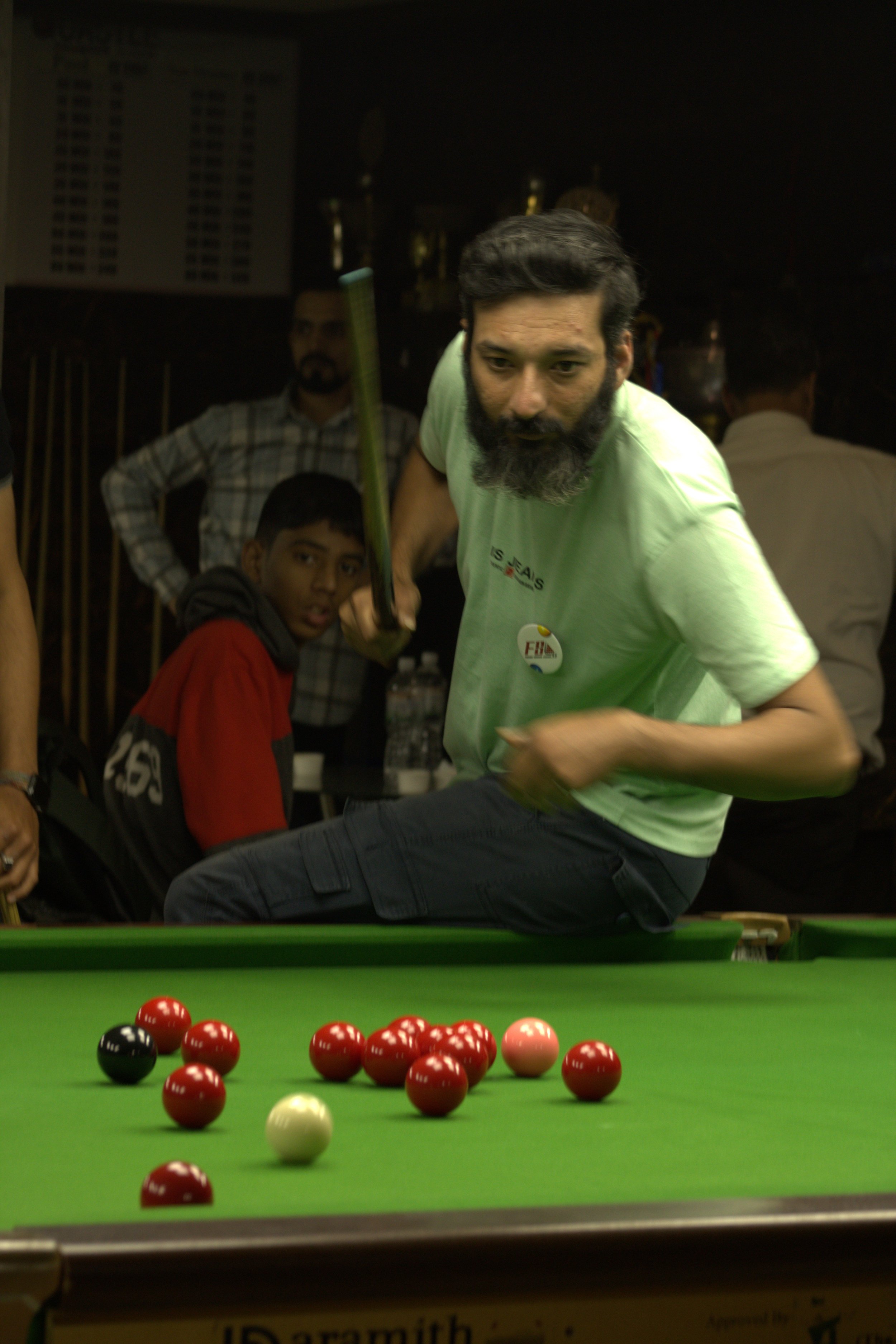 A man with a beard aiming a cue stick at a billiard pool game on a green table, with a boy and other people observing in the background.