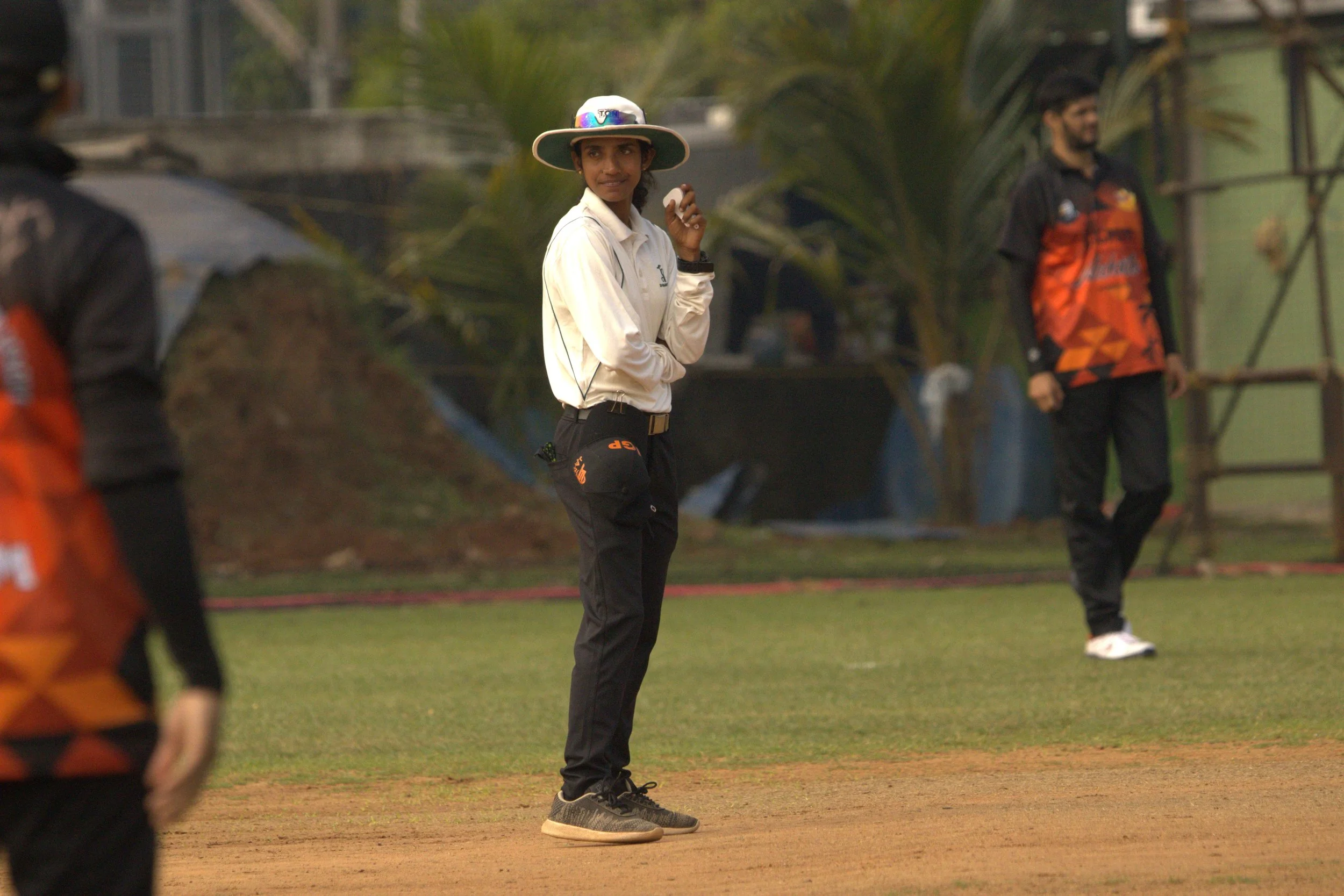 A woman in cricket uniform and wide-brimmed hat standing on a cricket field, holding a cricket ball in her right hand, with two other people in the background near a fence and trees.