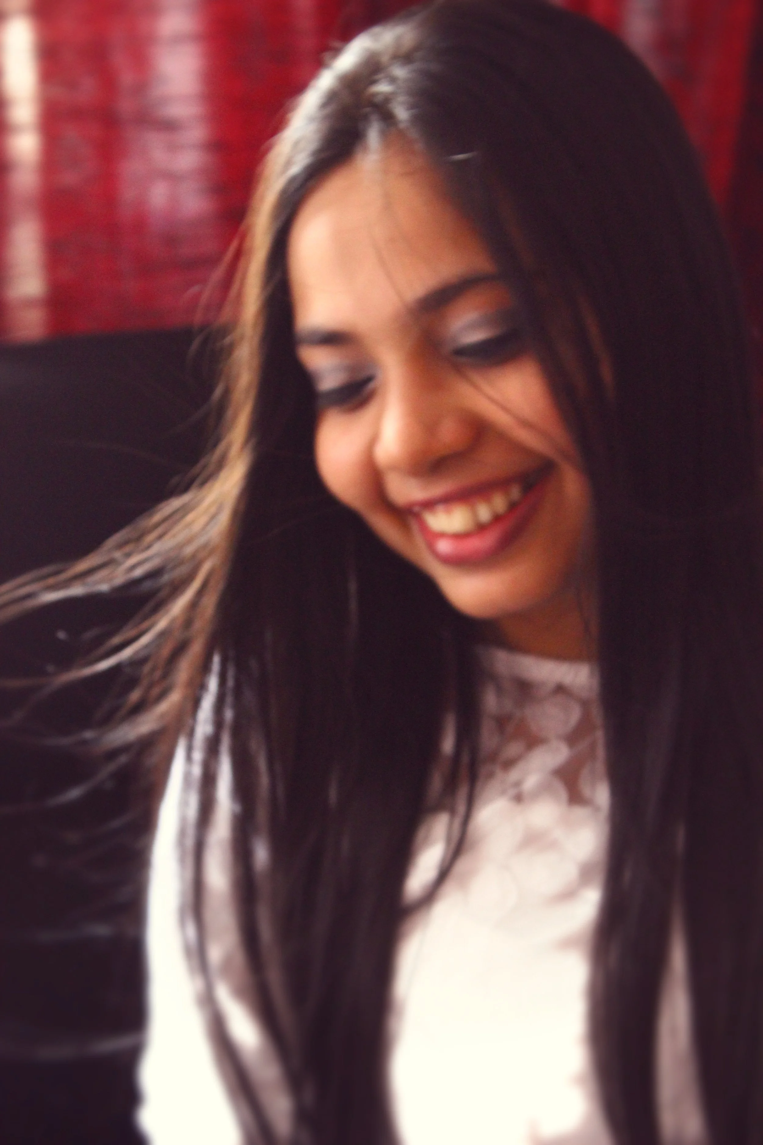 A woman with long dark hair smiling and looking downward, wearing a light-colored top, in front of a red background.