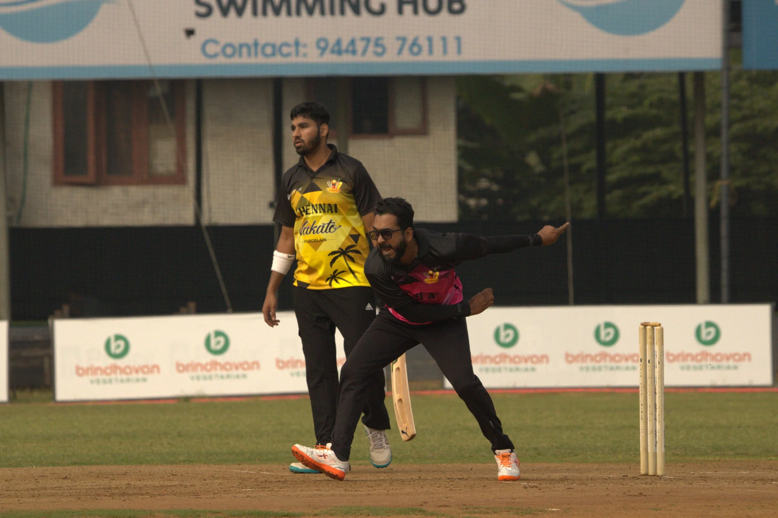 Two men playing cricket on a field, with one man in a pink and black uniform umpiring and focused on the game, and the other man in a yellow and black uniform standing nearby. There are advertising boards in the background.