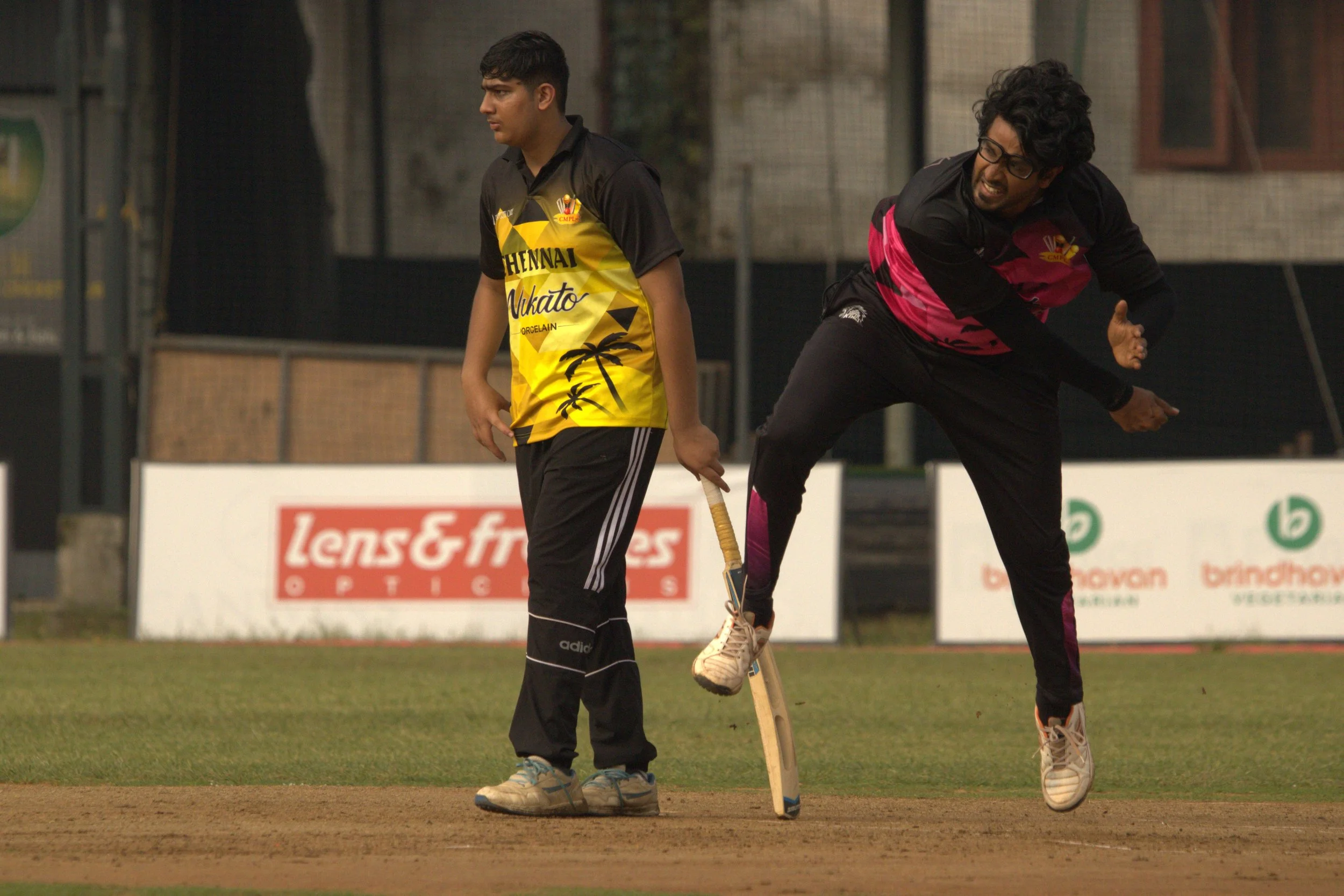 A young player in black and yellow sportswear holding a cricket bat and a coach or teammate in black and pink sportswear jumping in celebration on a cricket field.