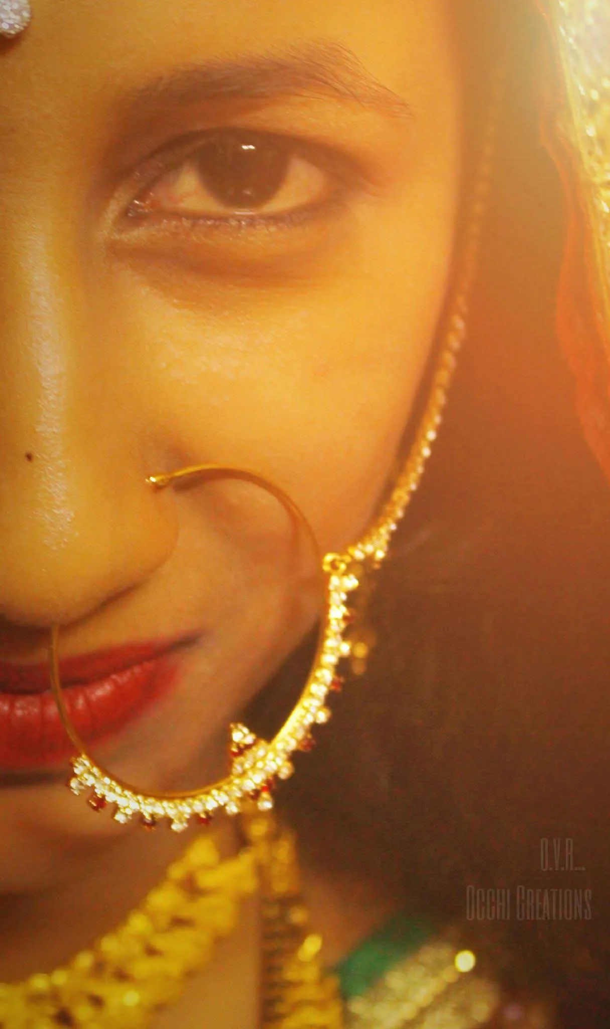 Close-up of a woman’s face with traditional jewelry, including large gold earrings, a nose ring, and a necklace, showing her eye, lips, and part of her forehead.