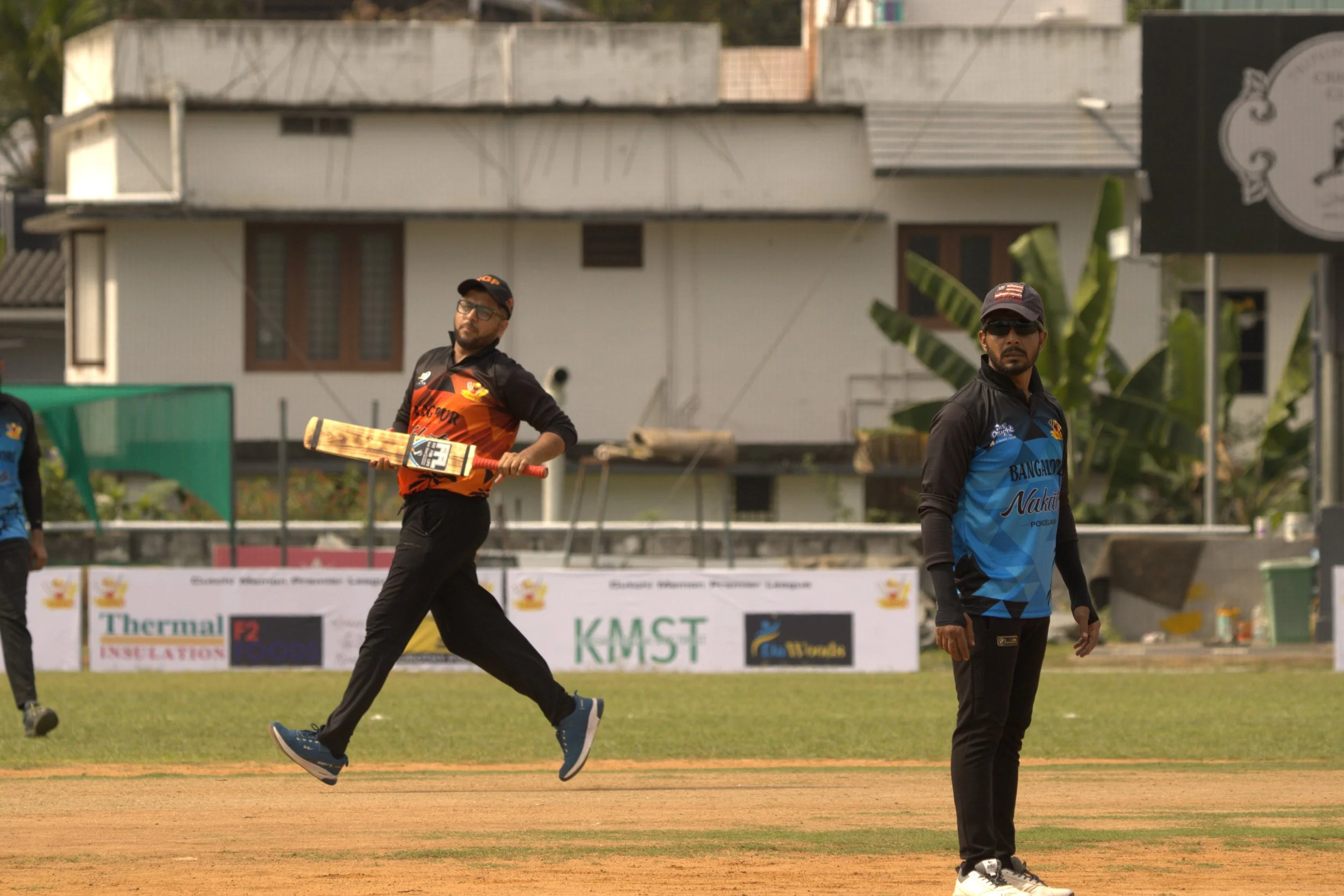 Two cricket players on the field, one holding a cricket bat and appearing to be celebrating, while the other stands nearby watching. The background features advertising banners and a building.