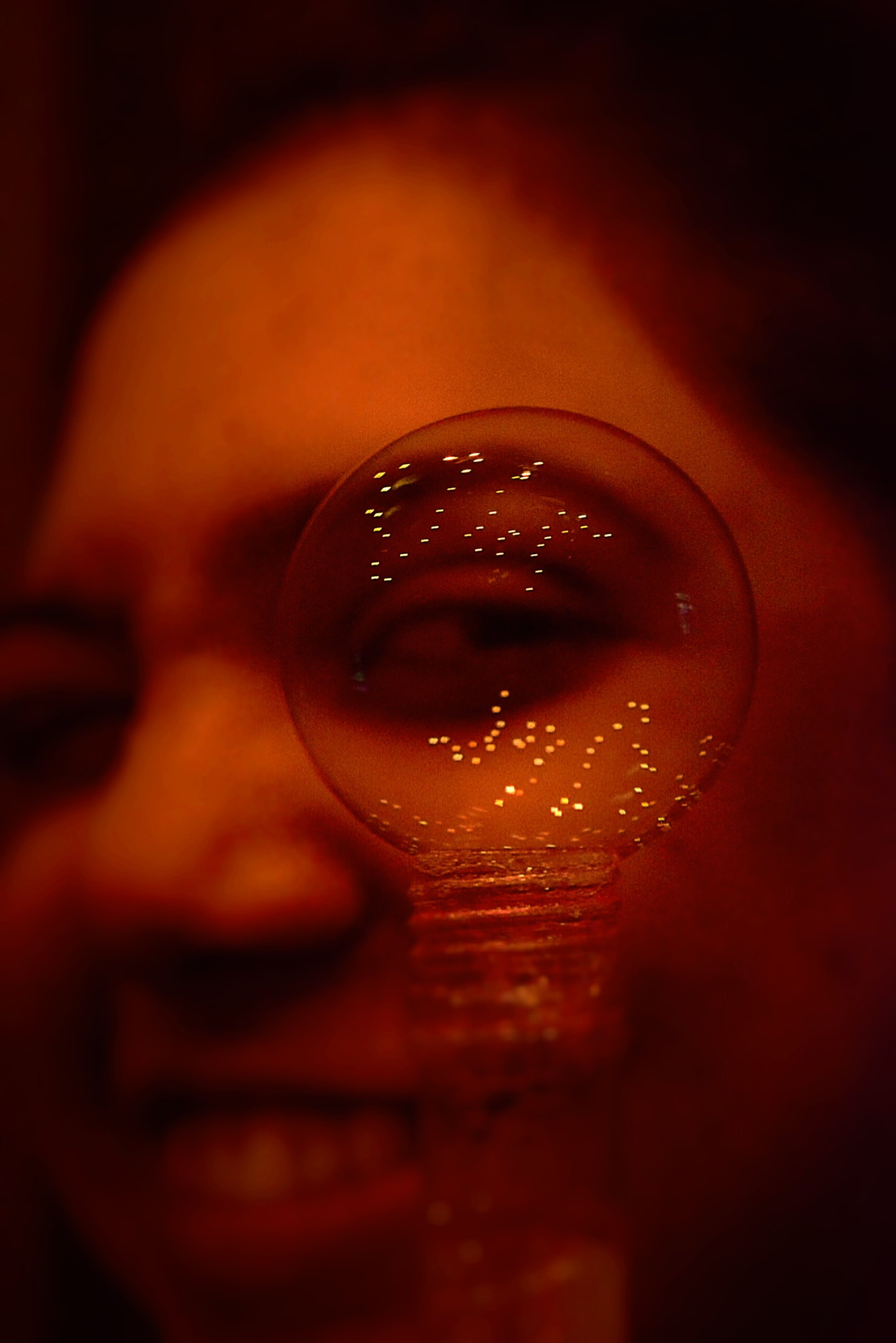 A person lying down with their head resting on a surface, looking through a bubble reflecting lights. The photo has a warm, reddish tone.