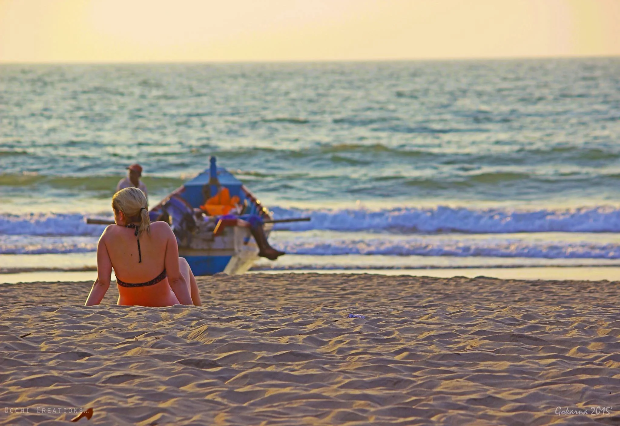 A woman sitting on the beach facing the ocean with a small boat and a man in the background during sunset.