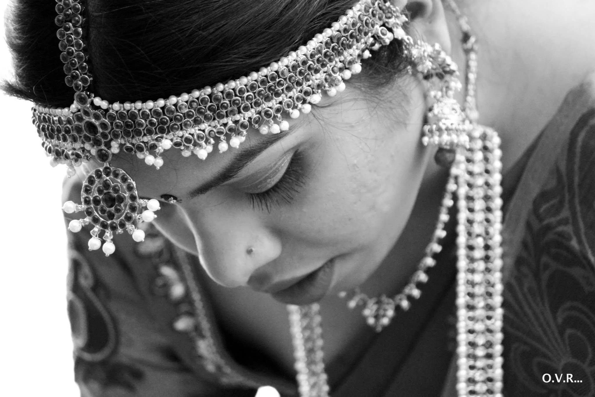 Close-up black and white photo of a woman wearing traditional jewelry, including a bejeweled headpiece, earrings, and necklace, with her eyes closed and head slightly bowed.