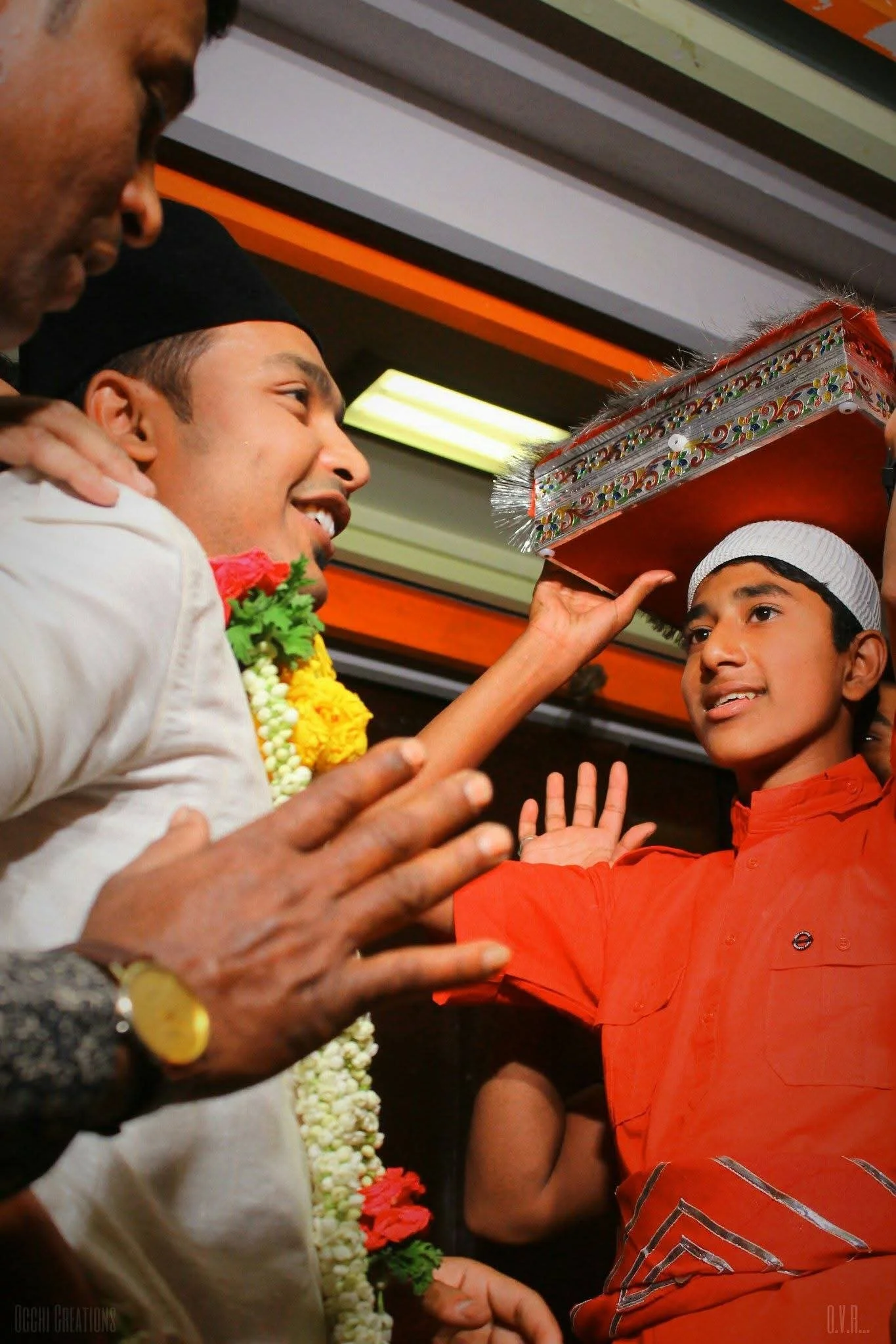 A young man in a red shirt and white cap holds a decorated box on his head during a celebration, while being blessed or receiving a ritual from a man wearing a garland of flowers, with other people gathered around.