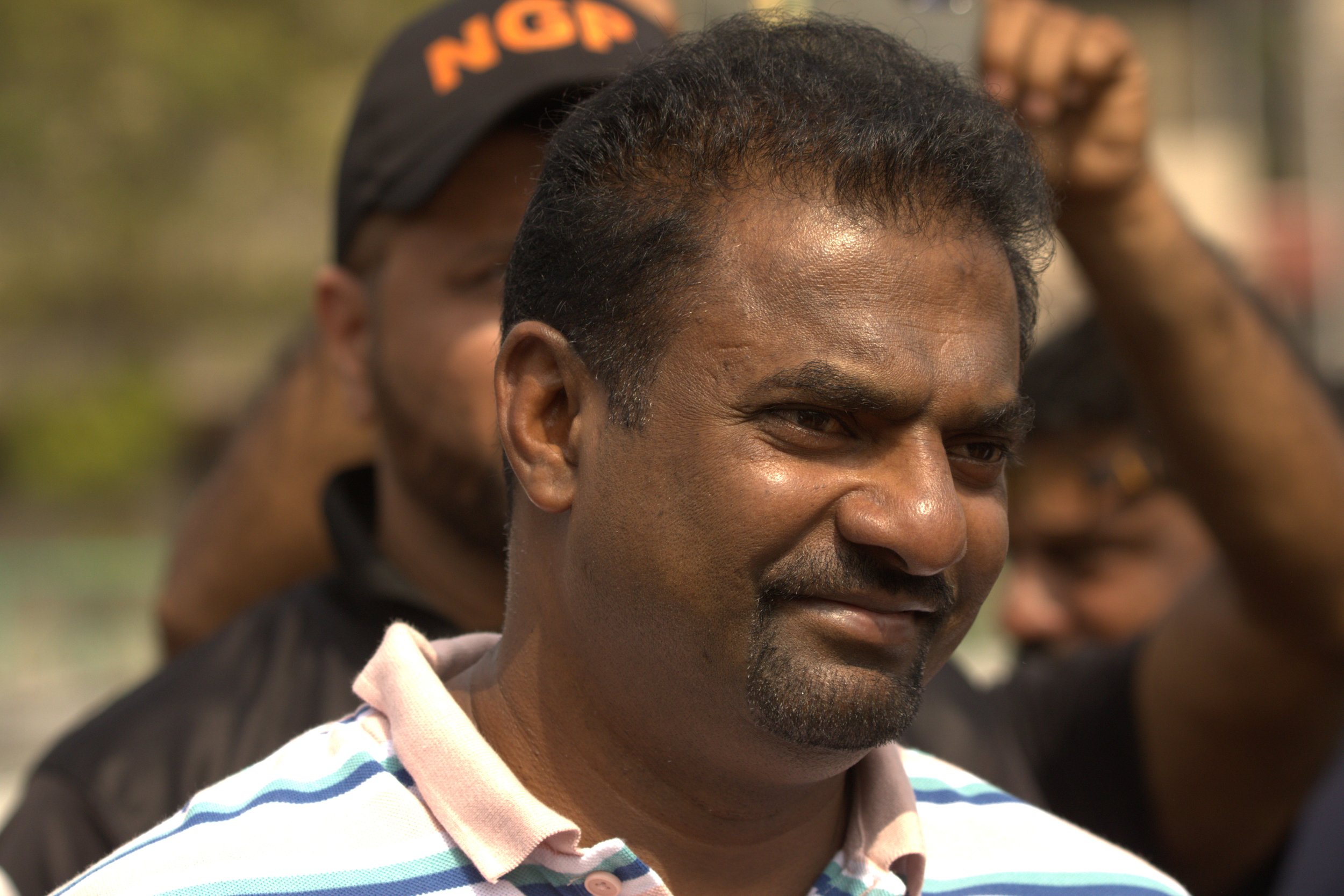 A man with dark hair, a mustache, and a goatee smiling, wearing a striped polo shirt, standing outdoors on a sunny day with blurred individuals in the background.